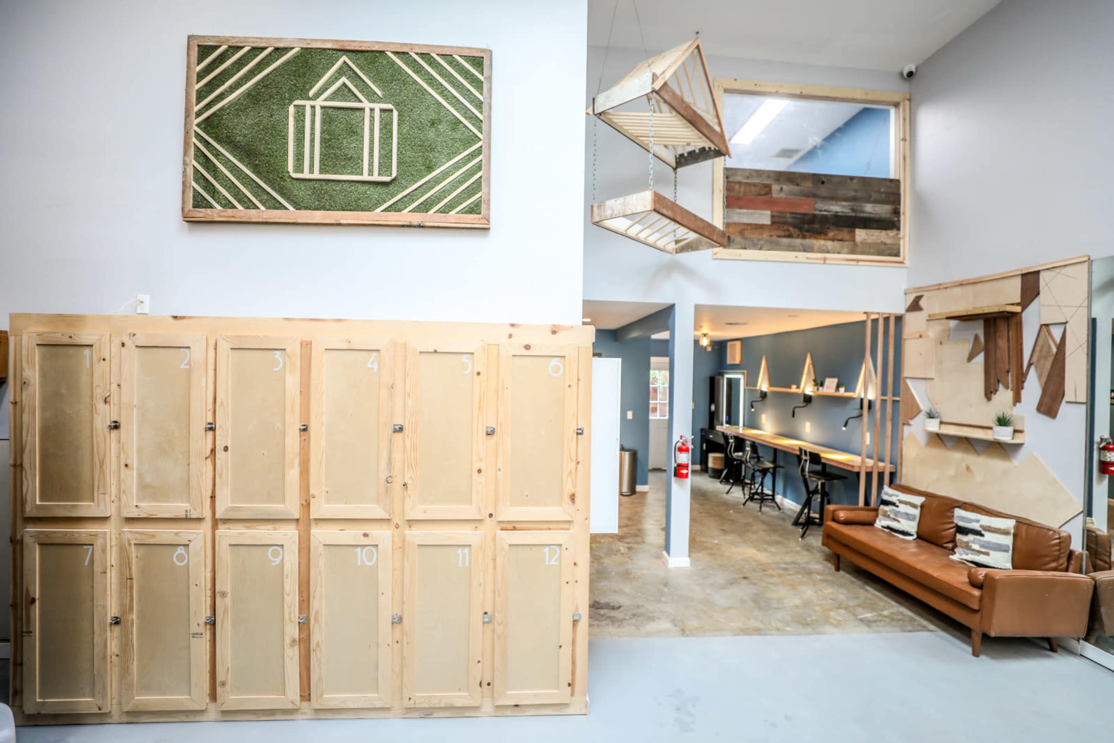 The image shows a modern interior space featuring wooden lockers, a minimalist seating area, and a second-story balcony overlooking a communal workspace.
