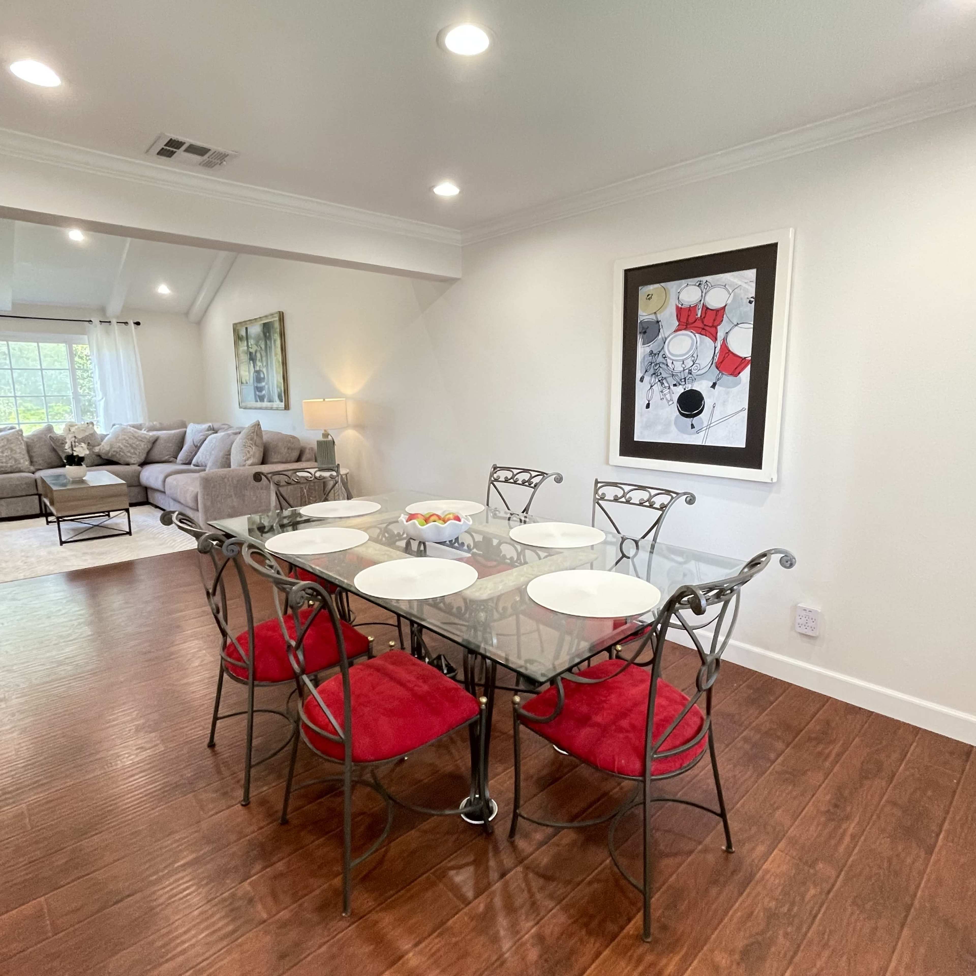 A glass dining table with red cushioned chairs is set in a brightly lit room featuring a large sofa and artwork on the wall.