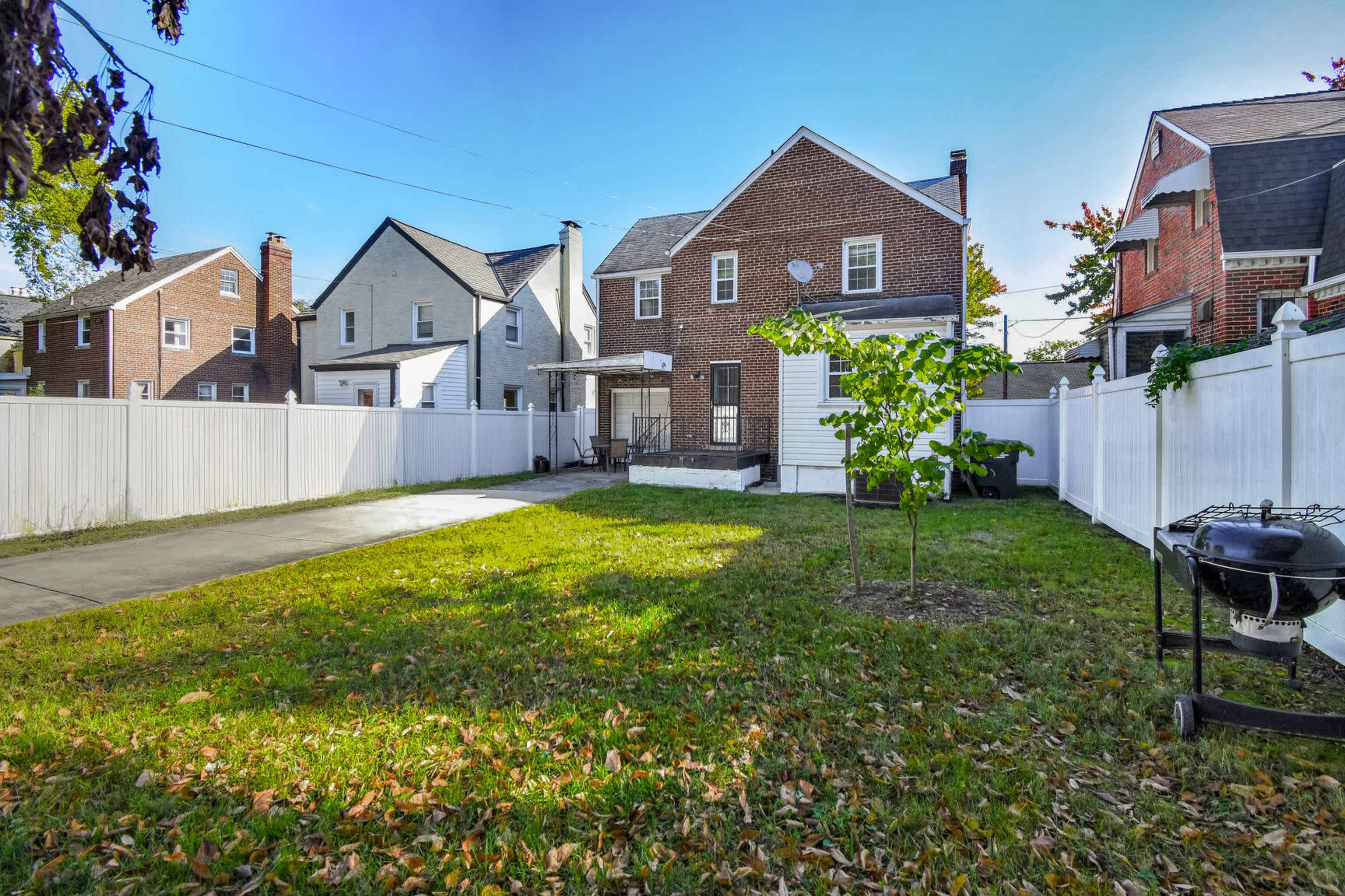 A backyard features a grassy area with a patio, surrounded by white fences and two houses in the background.