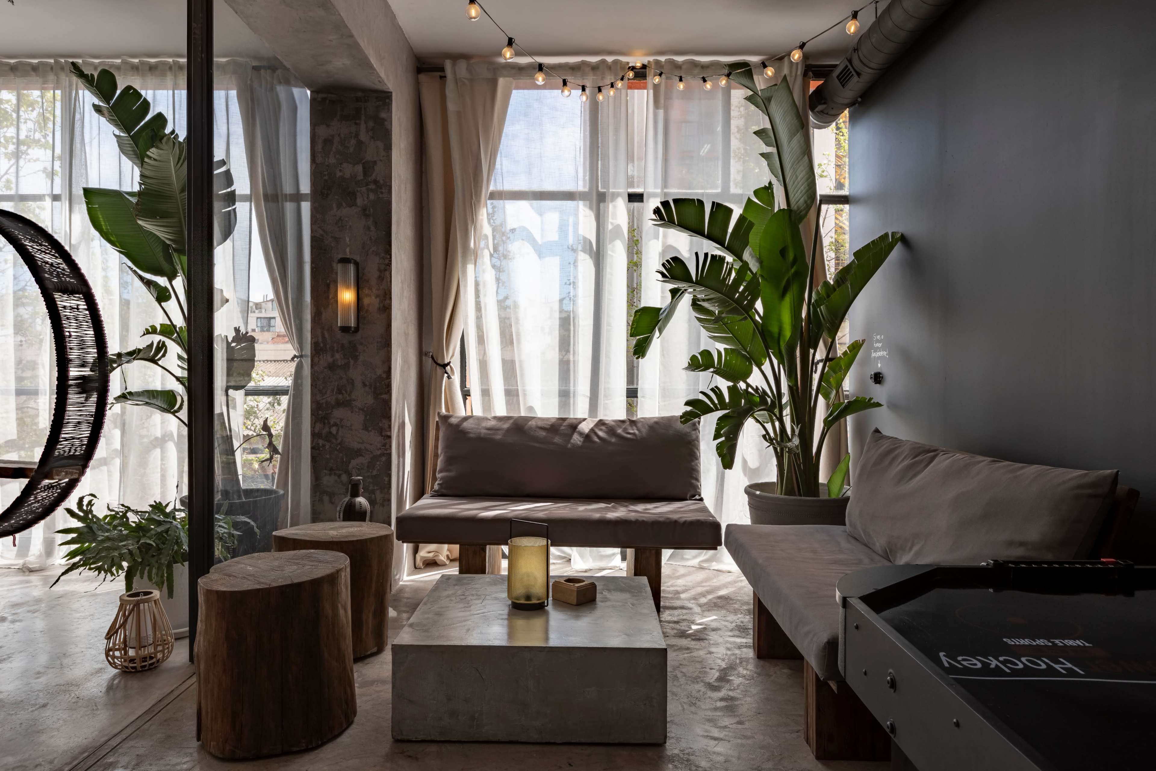A modern lounge area with a minimalist design featuring a gray sofa, wooden stools, indoor plants, and large windows covered with sheer curtains.