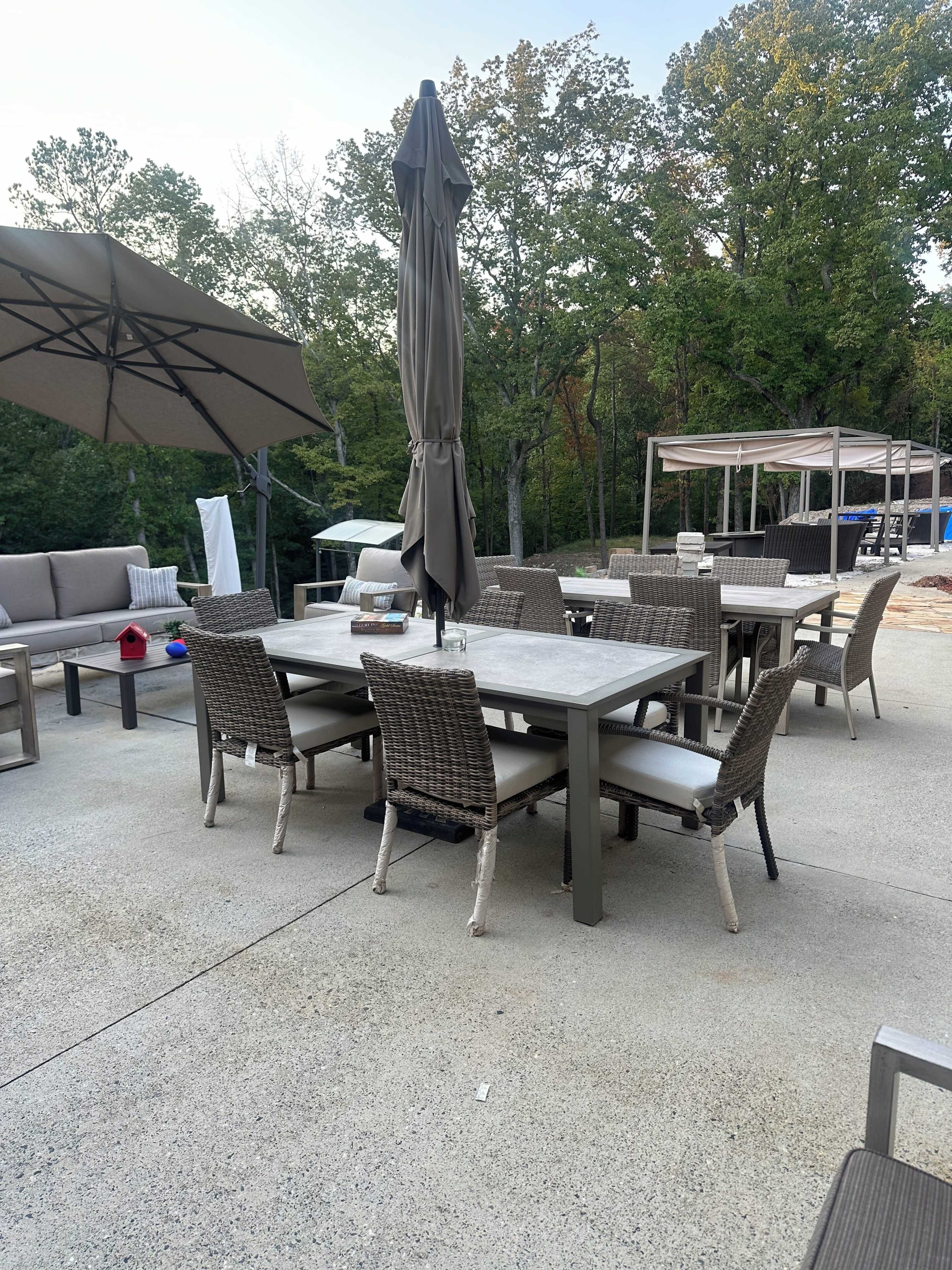 A patio area features a dining table surrounded by wicker chairs, an umbrella, and lounge seating amidst trees in the background.