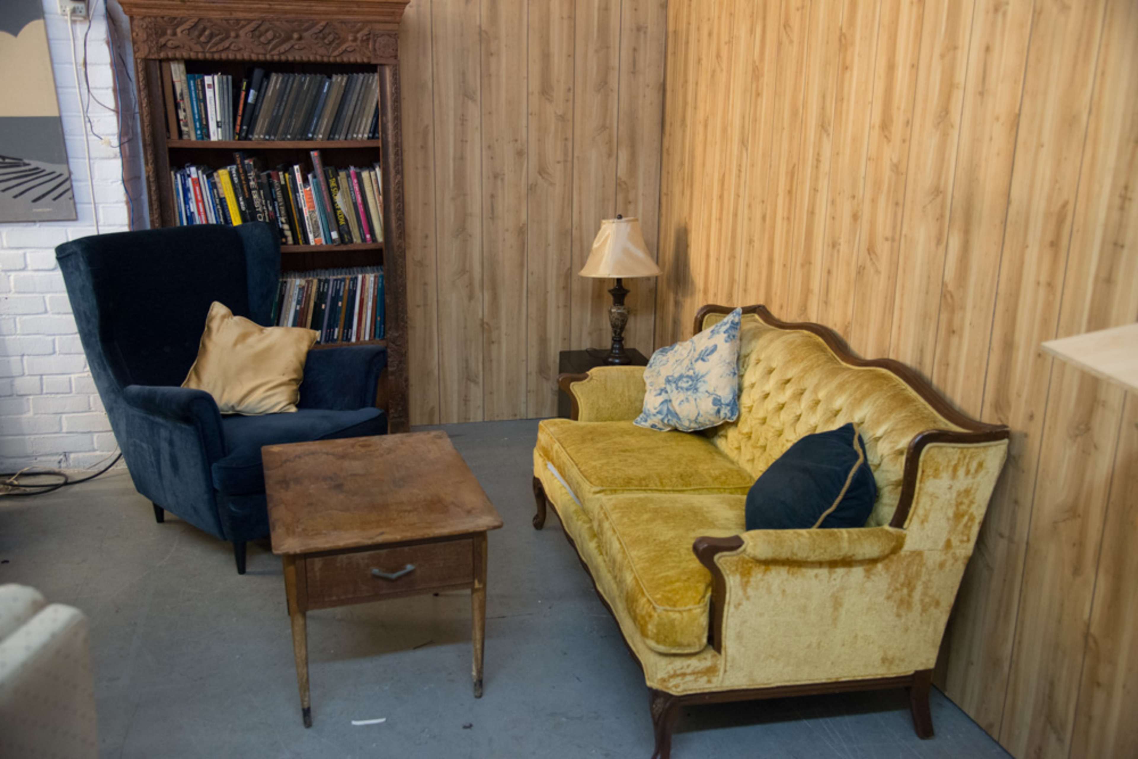 A small living area with a blue armchair, a yellow tufted sofa, a wooden coffee table, and a lamp next to a bookshelf against a wooden-paneled wall.