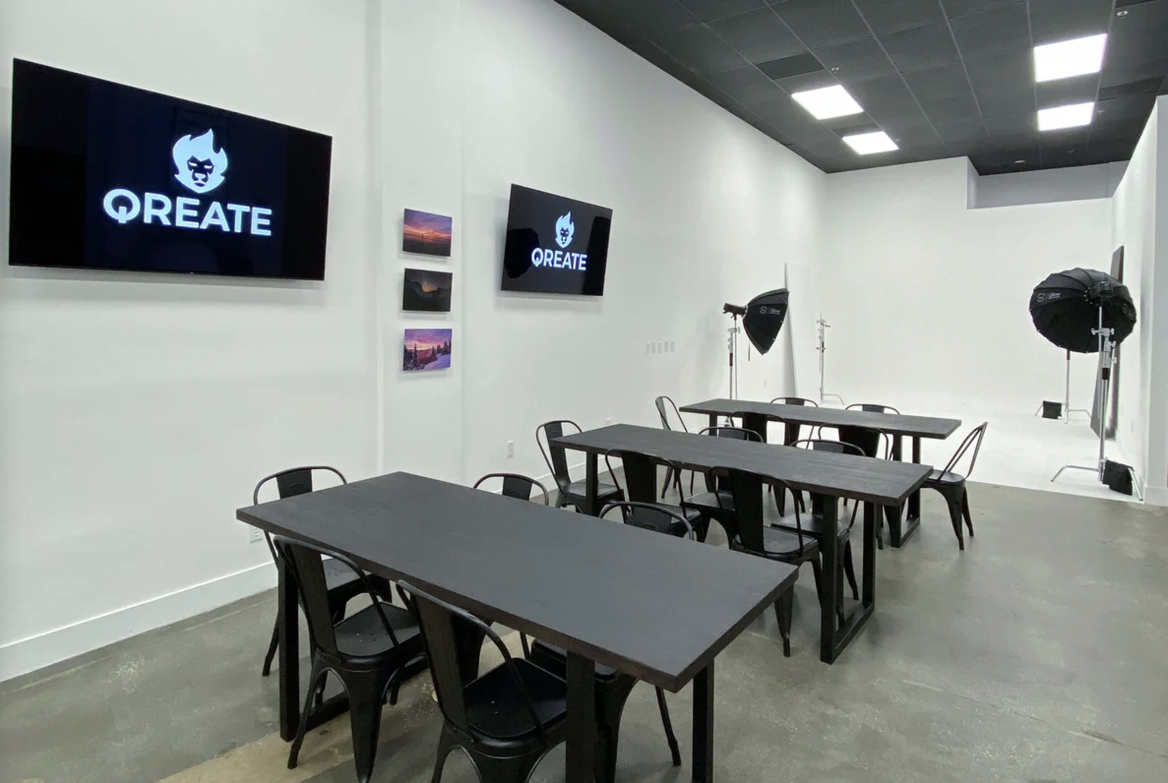 A spacious, white-walled room with multiple black tables and chairs, two monitors displaying the logo "QREATE," and studio lighting equipment in the background.