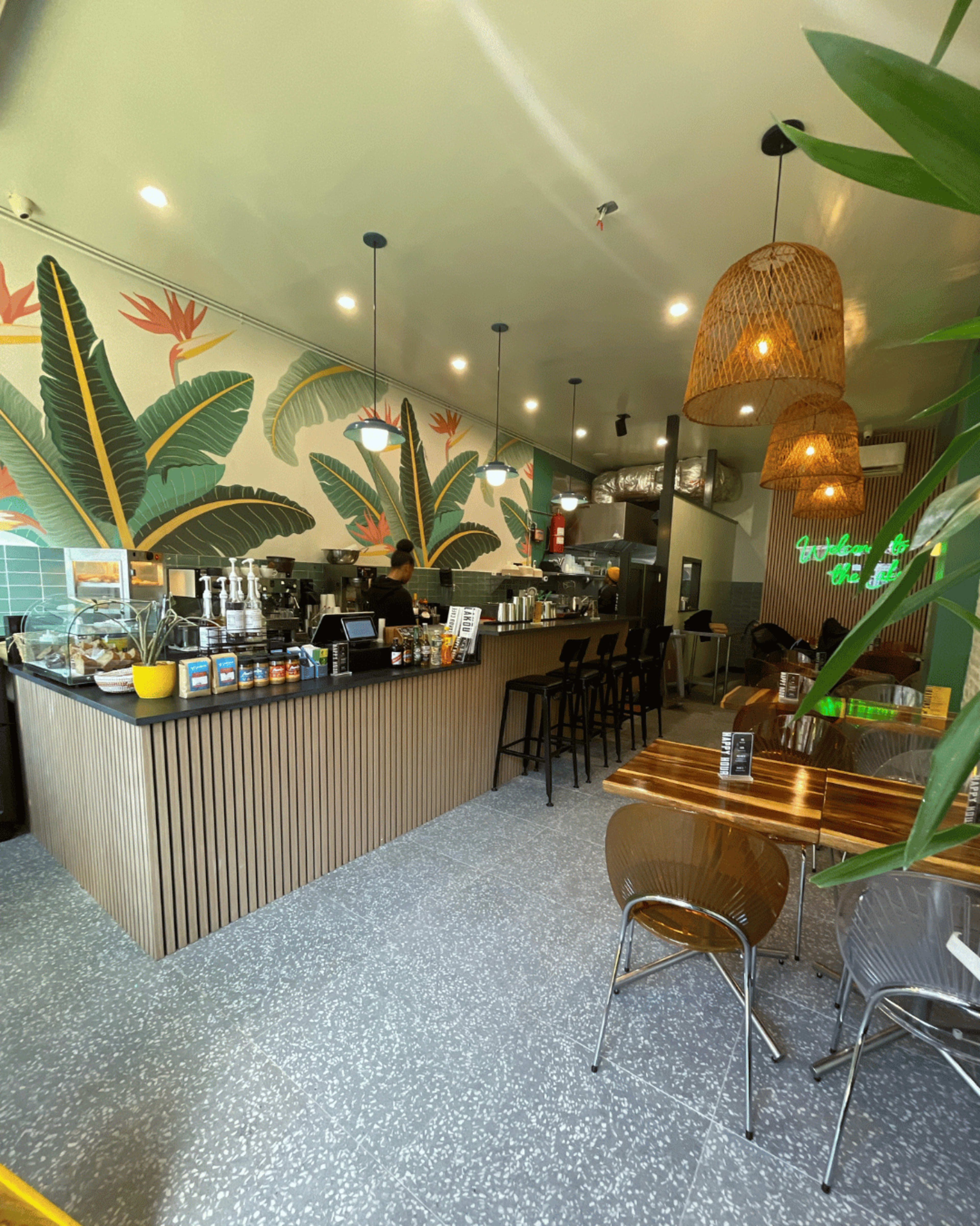 The image shows a modern café interior with a wooden counter, bar stools, and tropical-themed wallpaper.