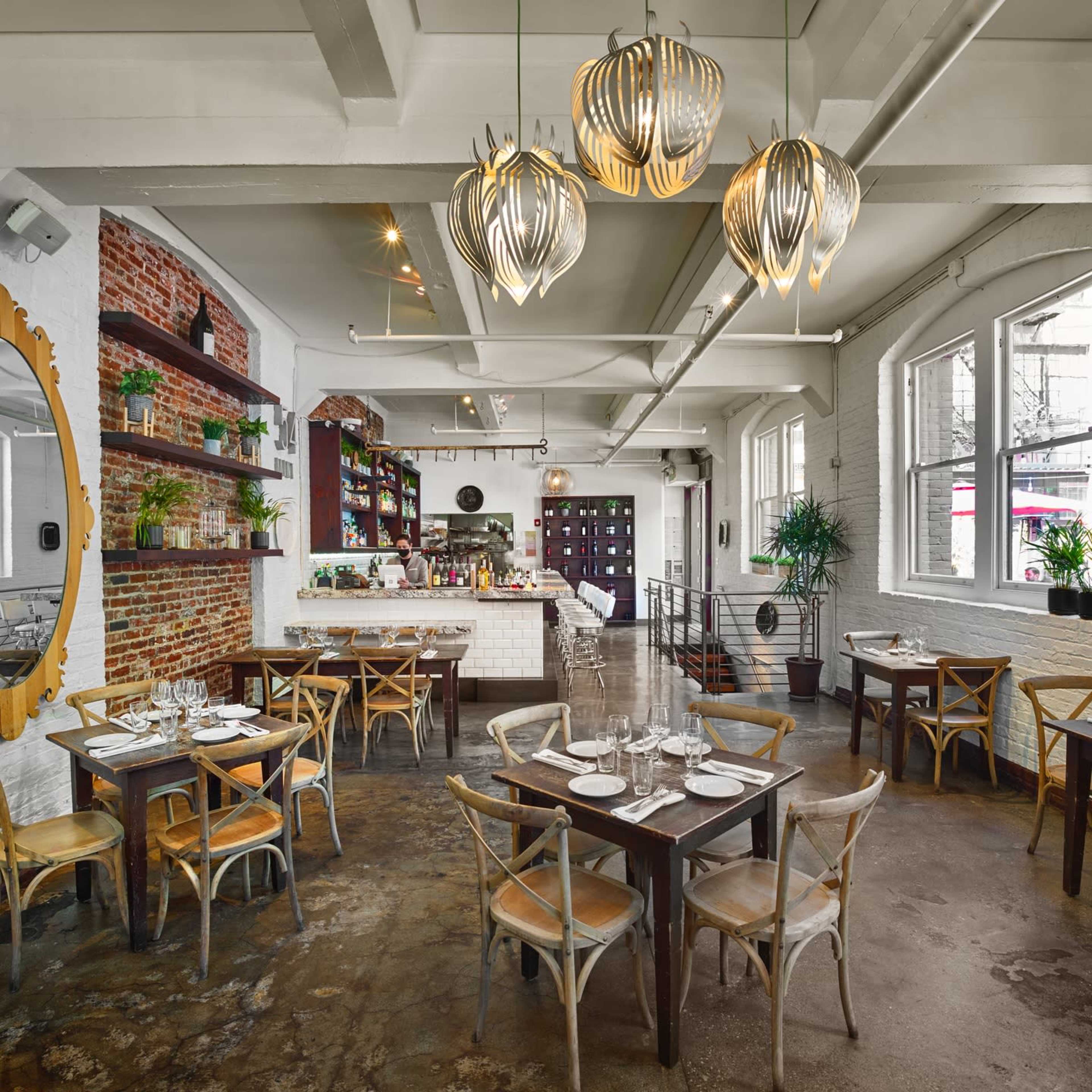 The image shows a restaurant interior with wooden tables, hanging light fixtures, and a mix of brick and white walls, featuring plants and a bar area.