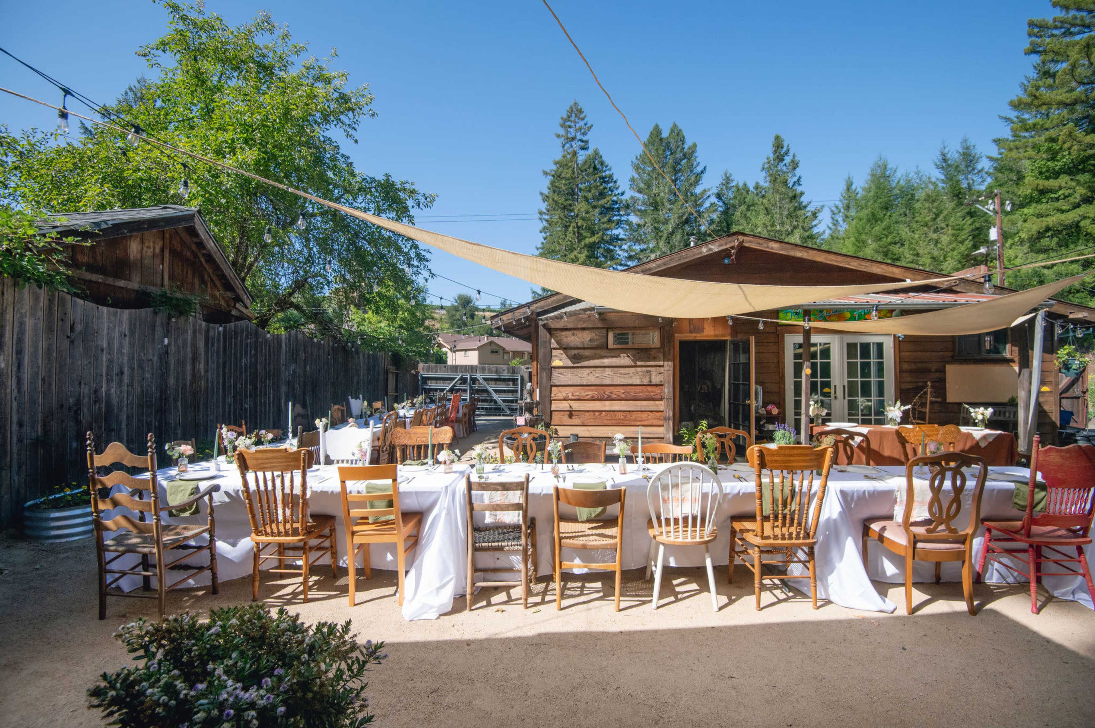 A long table set for an outdoor dining event is arranged in front of a rustic cabin surrounded by trees.