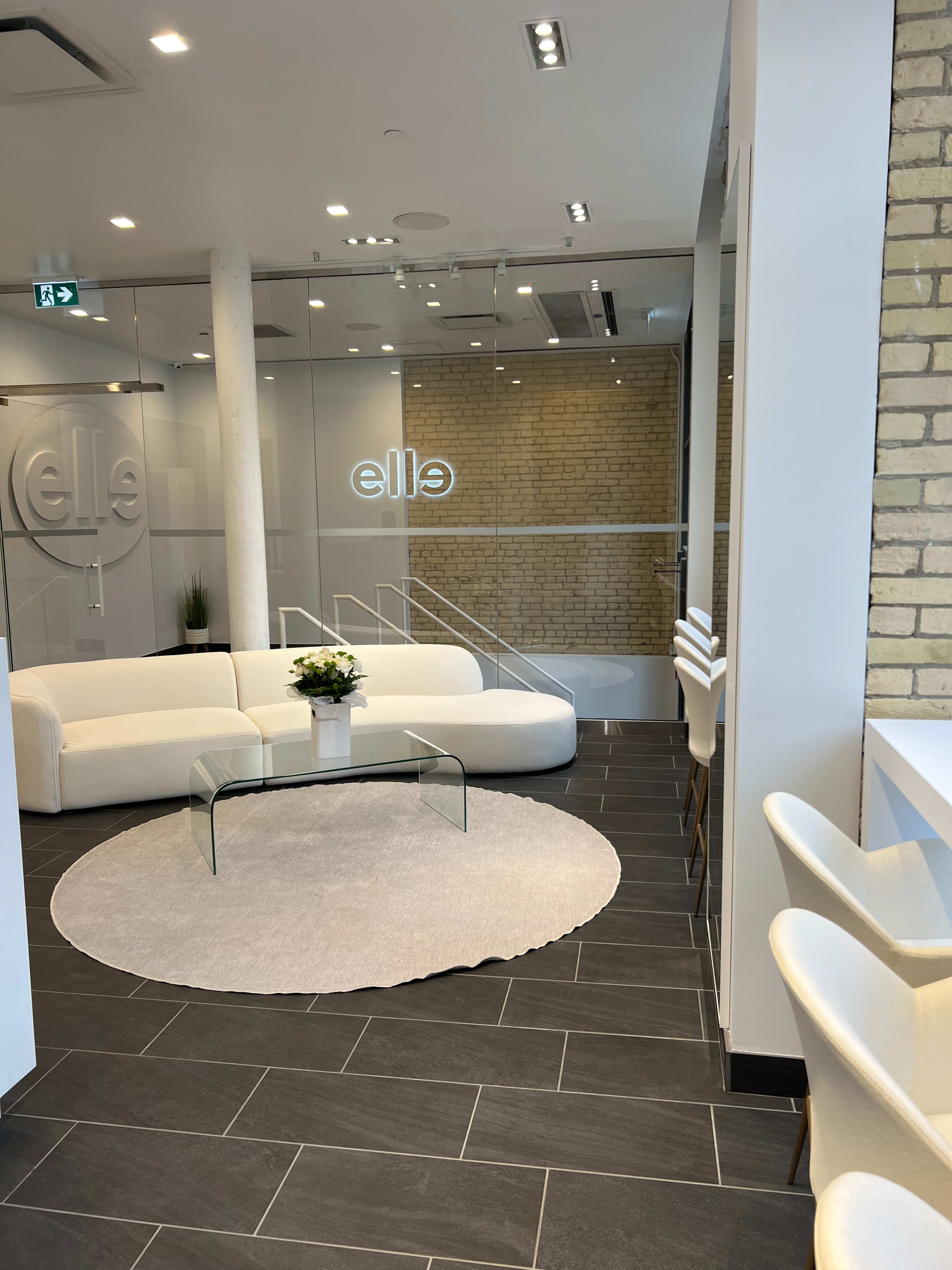 A modern, minimalist reception area featuring a white sofa, a glass coffee table, and a rug on a dark tiled floor, with a brick wall and an illuminated sign reading "elle" in the background.