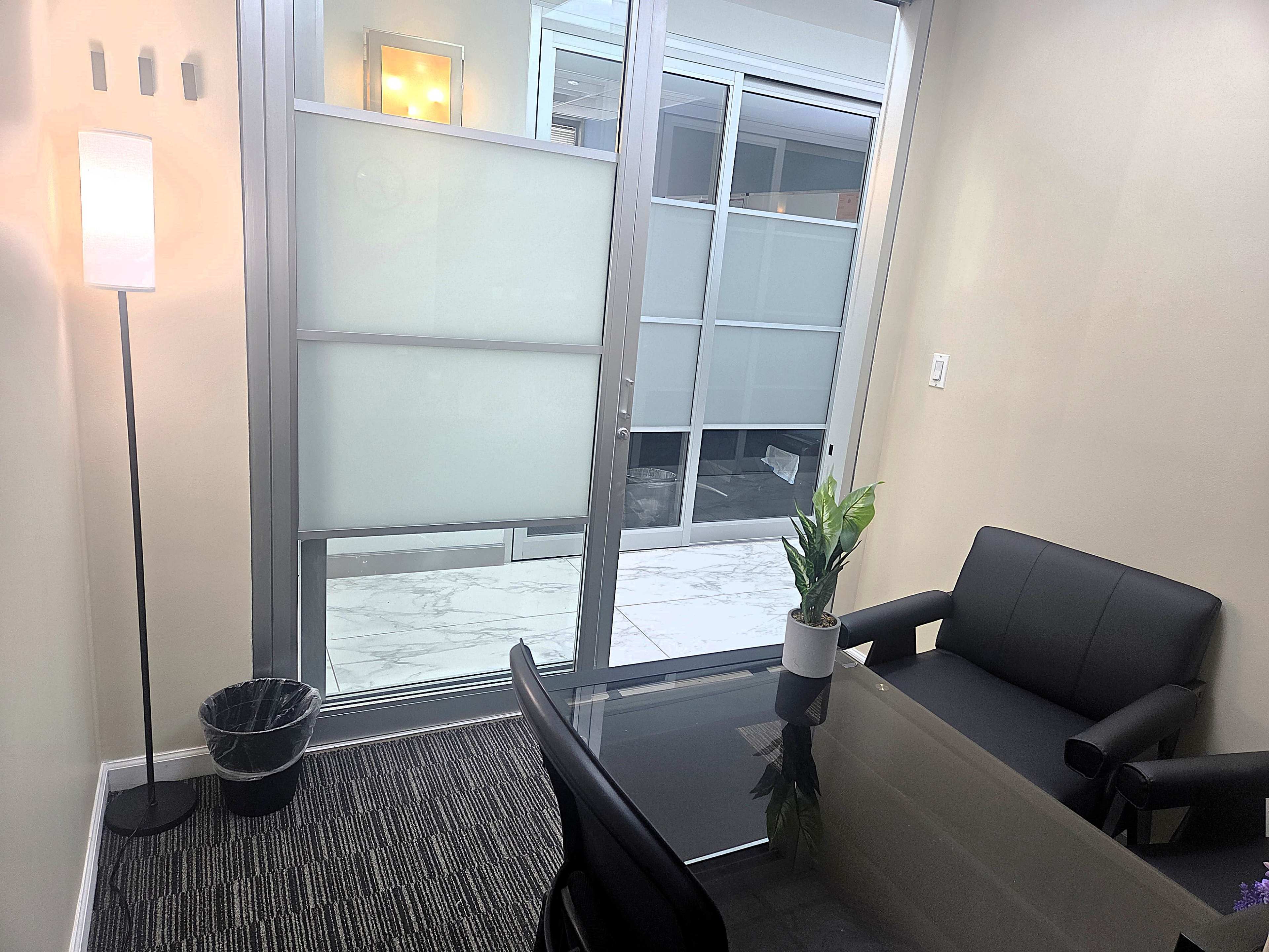 The image shows a small office space featuring a glass door, a black seating area, a potted plant, and a light-colored wall.