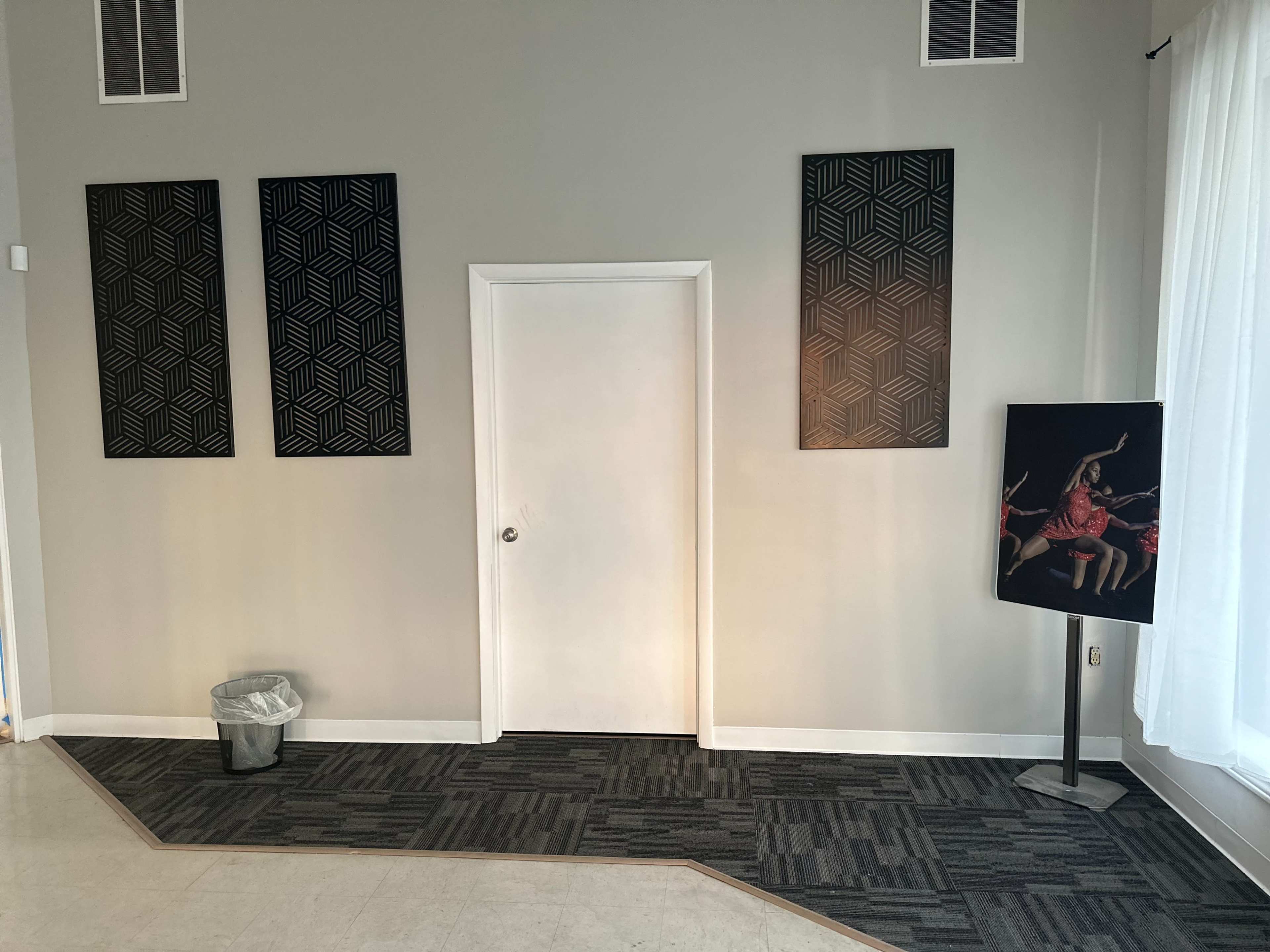 The image shows a minimalist interior with three patterned wall panels, a closed white door, a standing display featuring a dancer, and a trash can on the floor.