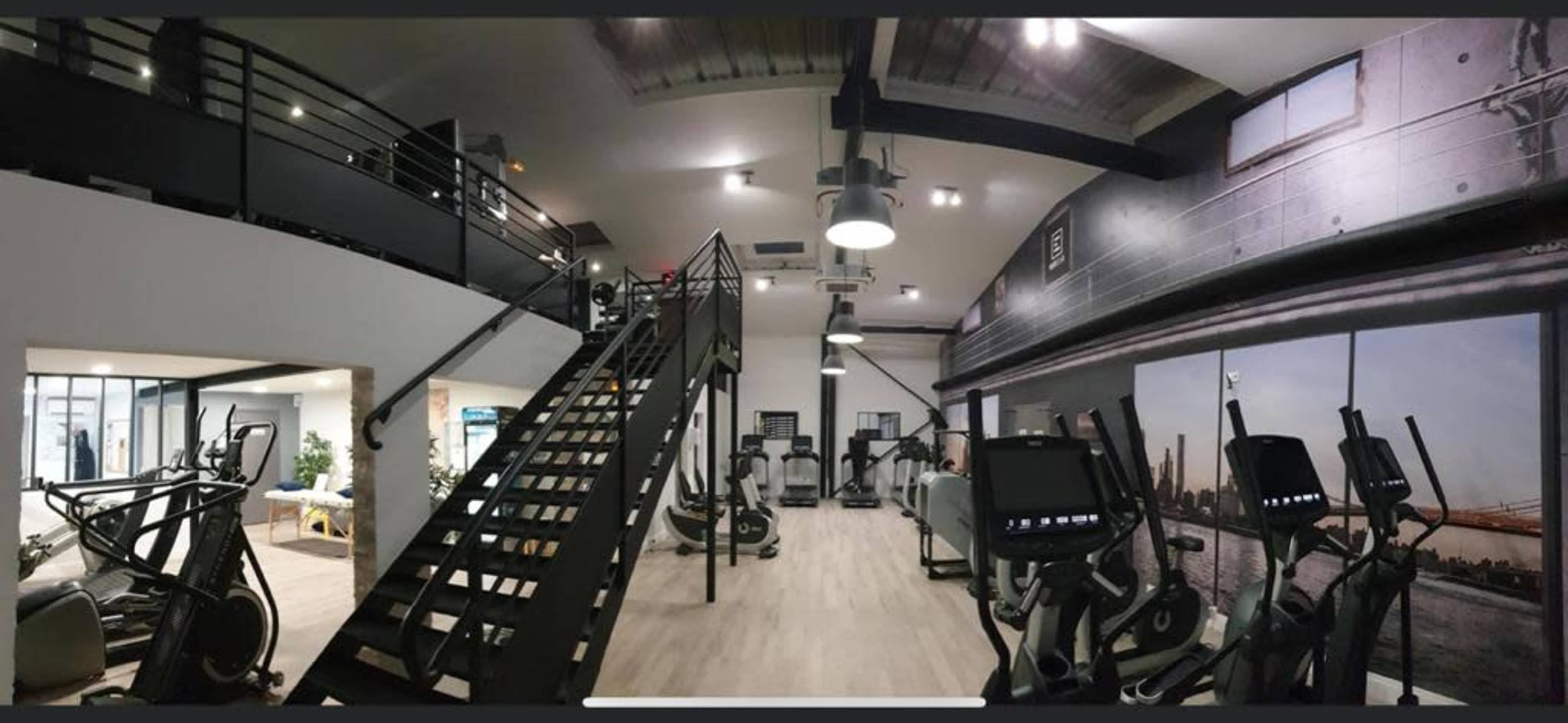 A modern gym interior featuring various exercise machines, a staircase, and large windows that let in natural light.