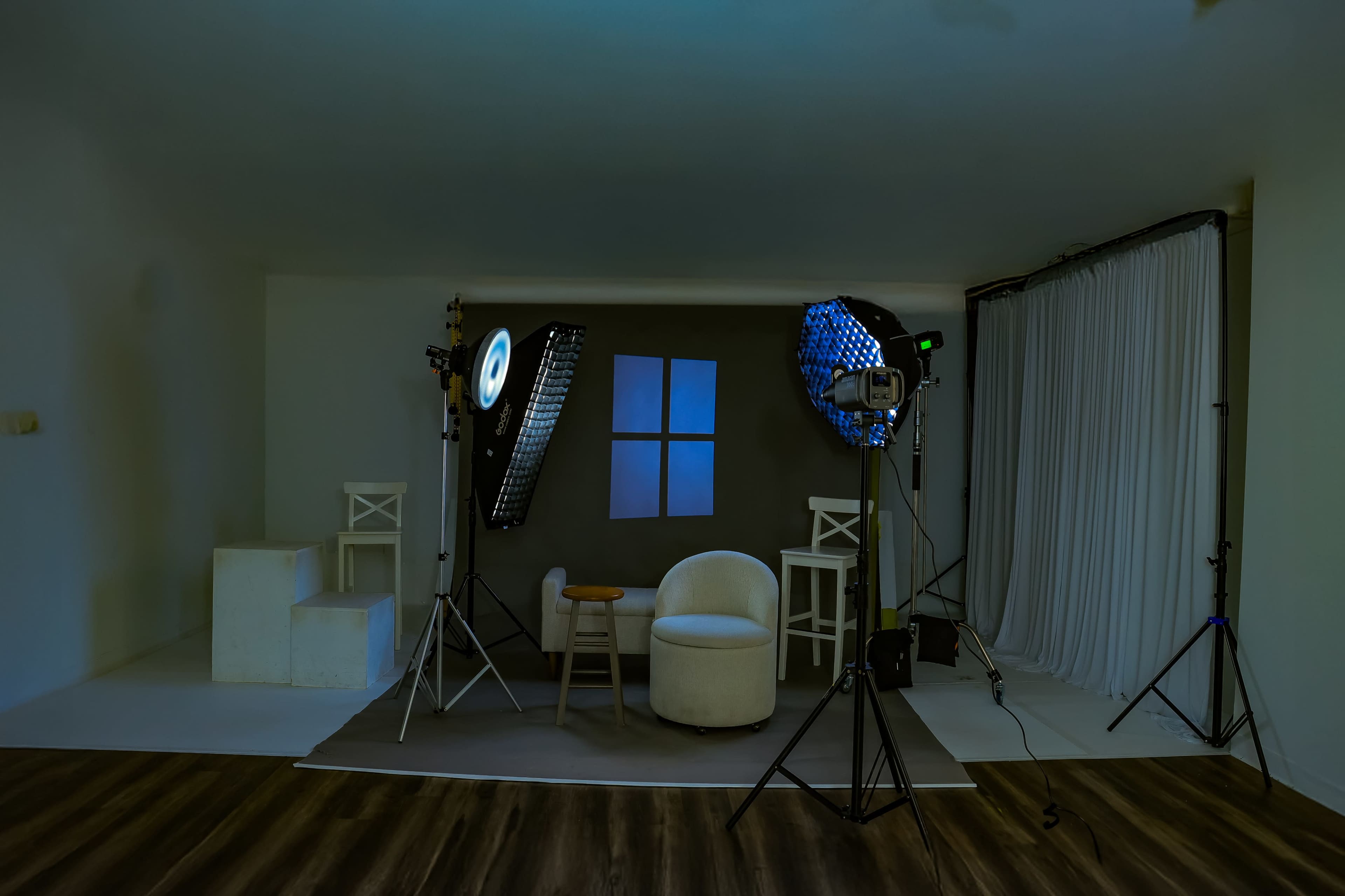 The image shows a photography studio setup with a white backdrop, lighting equipment, and a round chair arranged in front of a window painted on the wall.