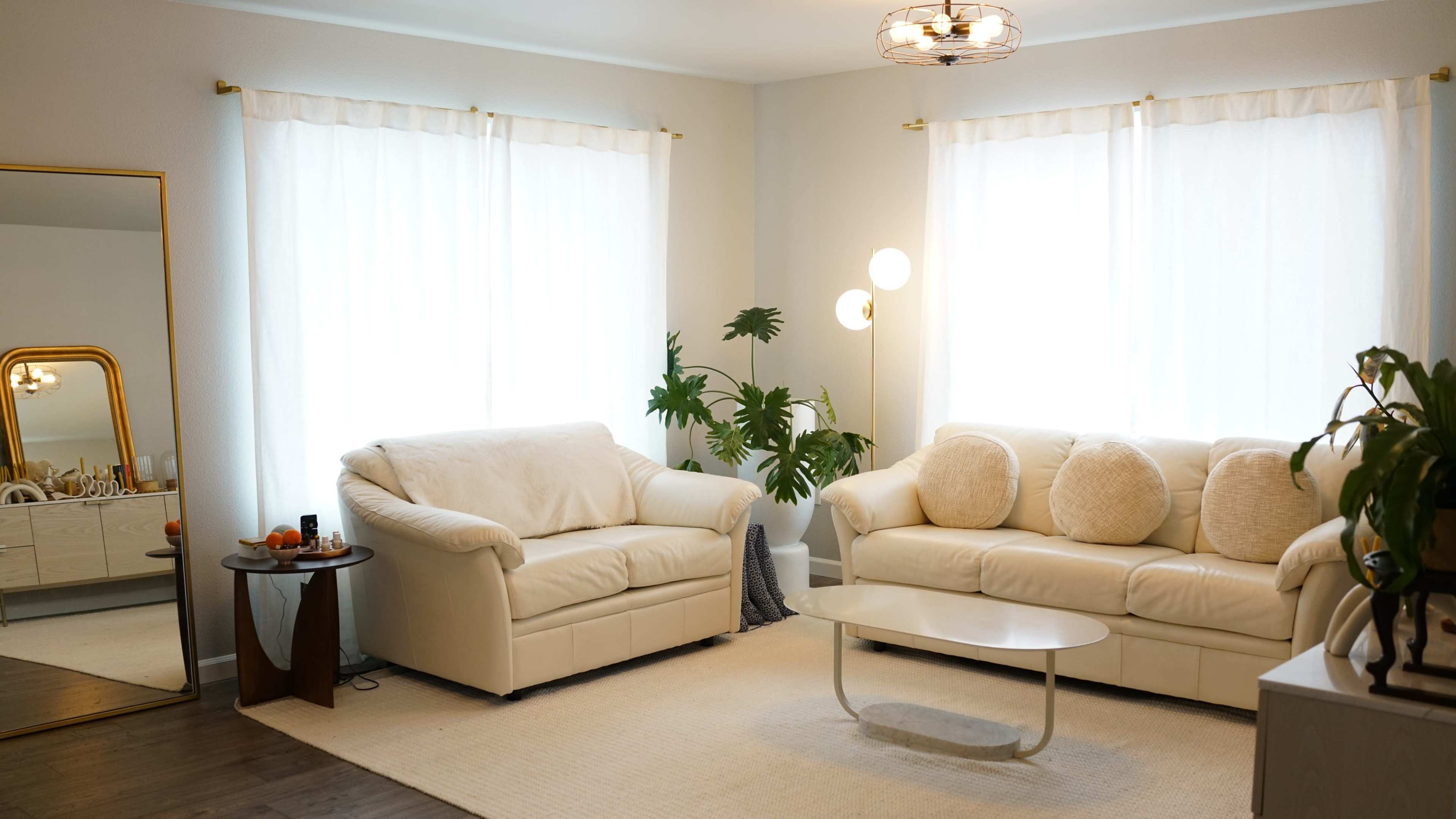 A living room with two white sofas, a round coffee table, a large mirror, and a potted plant near two windows with sheer curtains.