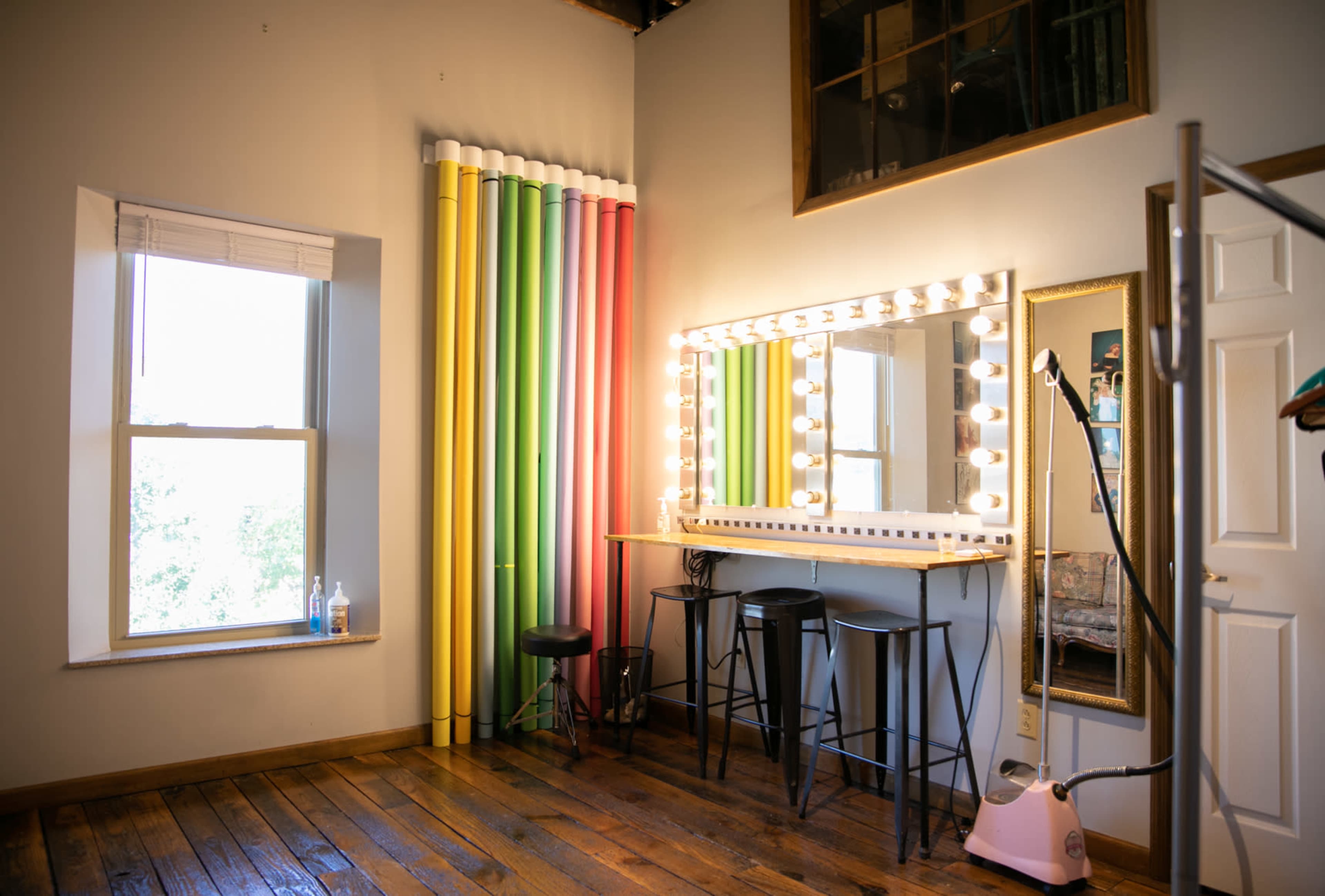 A brightly lit makeup area with a large mirror surrounded by bulbs, a wooden floor, and colorful backdrop rolls.