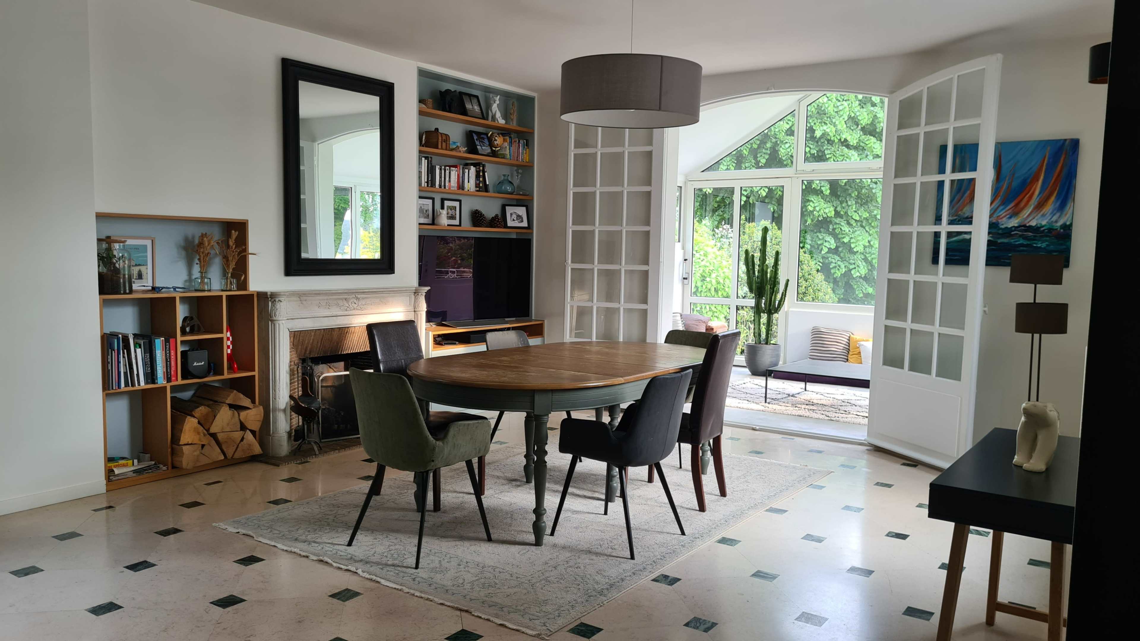 A round wooden dining table surrounded by upholstered chairs is set in a room with a fireplace, bookshelves, and large windows opening to a patio with greenery.