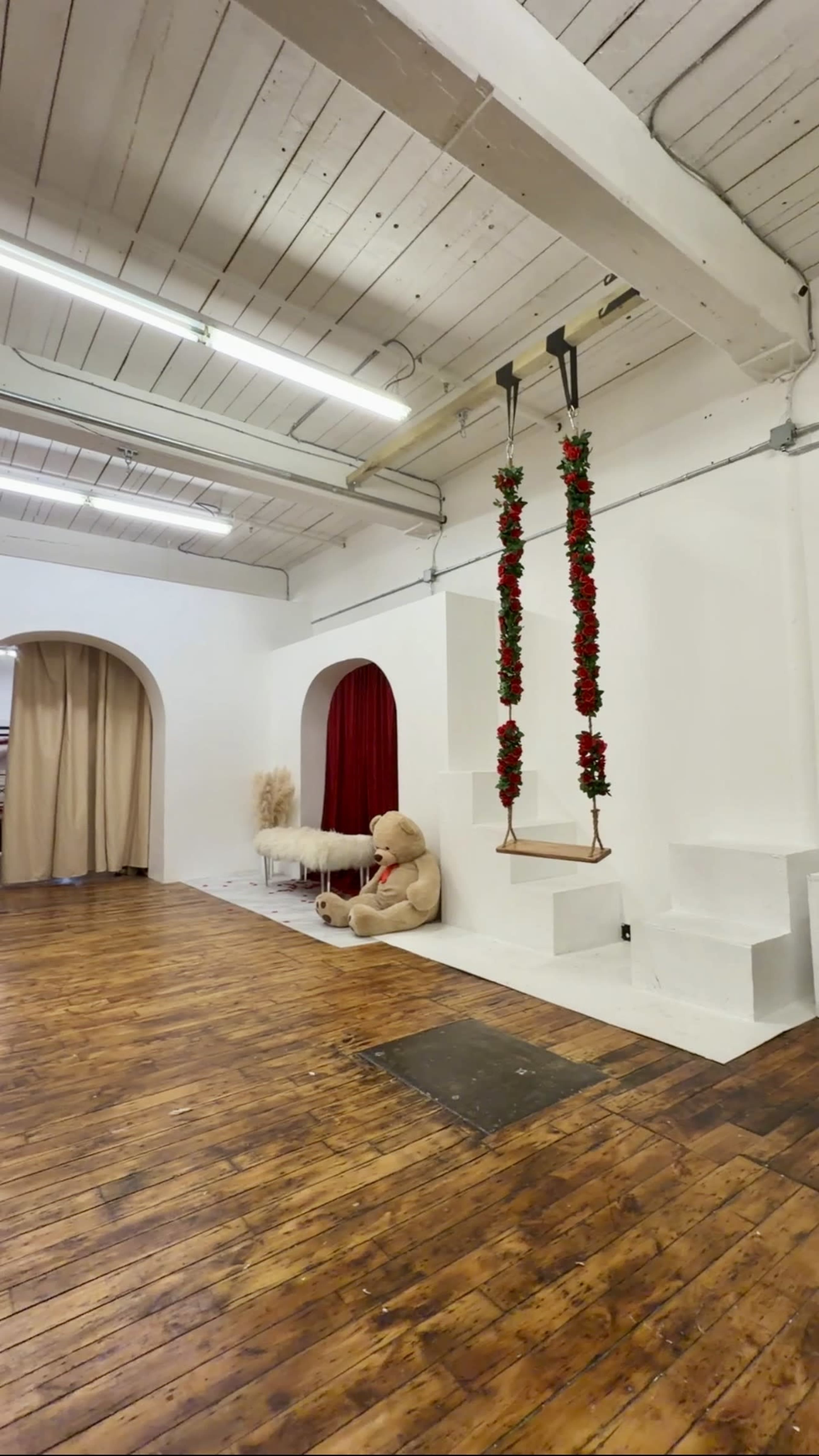 The image shows a spacious room with wood flooring, featuring a large teddy bear, a swing decorated with red flowers, and arched openings leading to curtained areas.