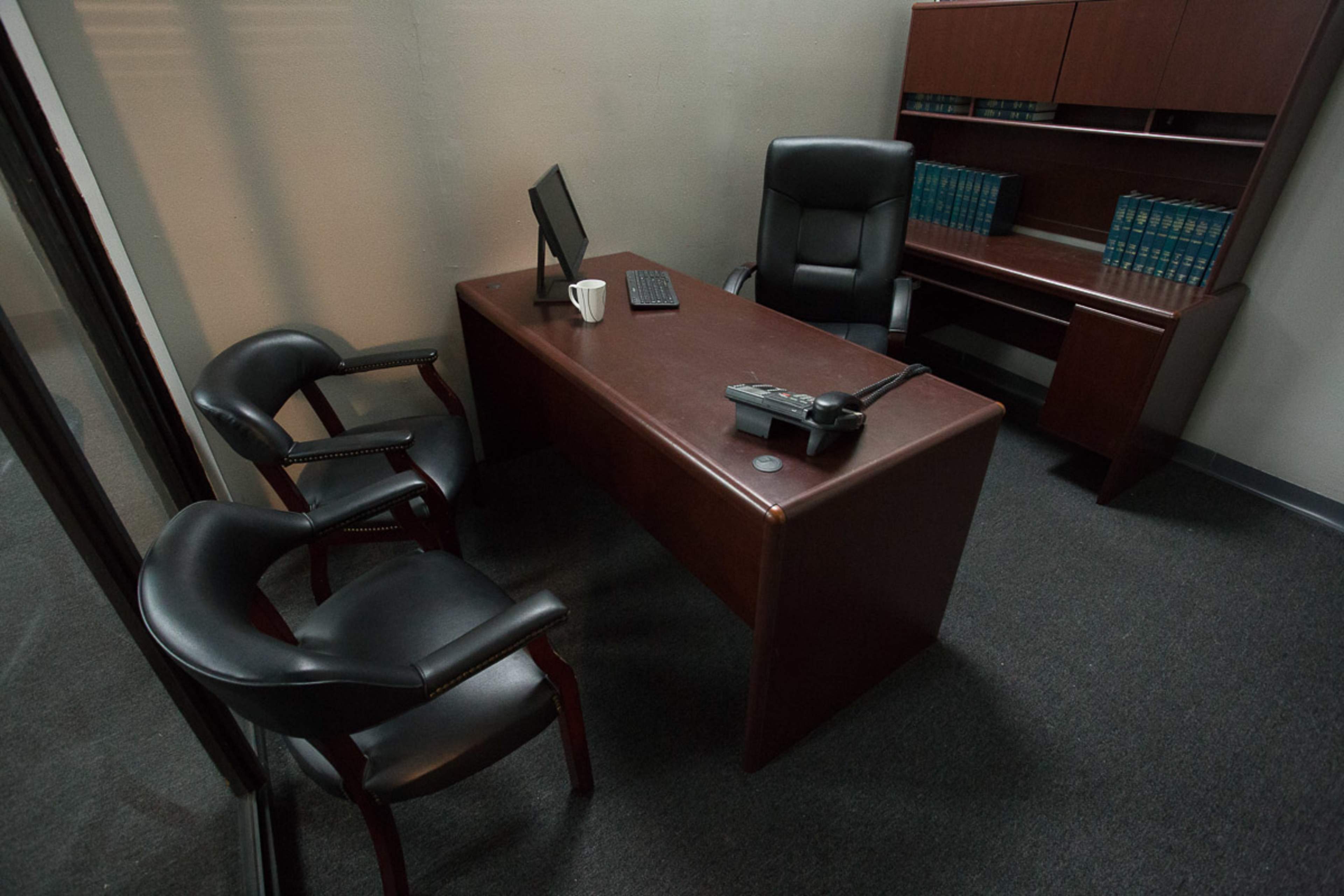 A minimalist office space features a wooden desk, a computer monitor, two leather chairs, and a coffee mug.