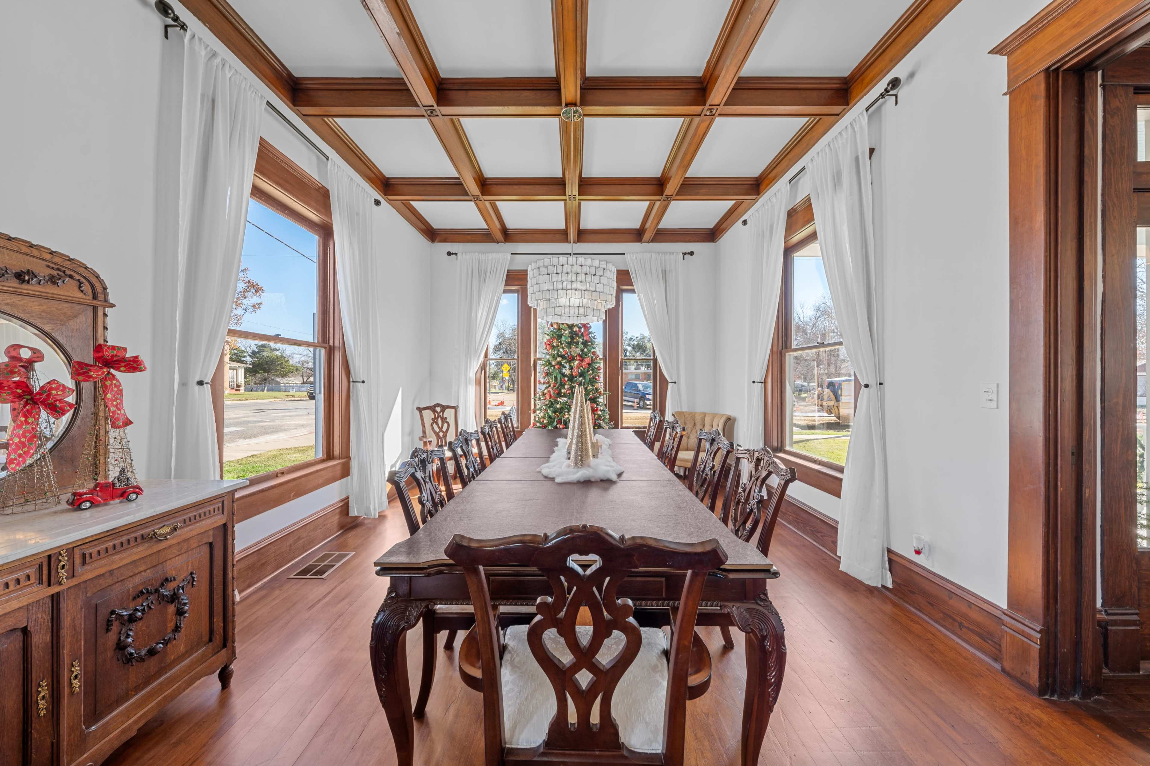 A long dining table with wooden chairs is set in a room featuring large windows and a coffered ceiling.