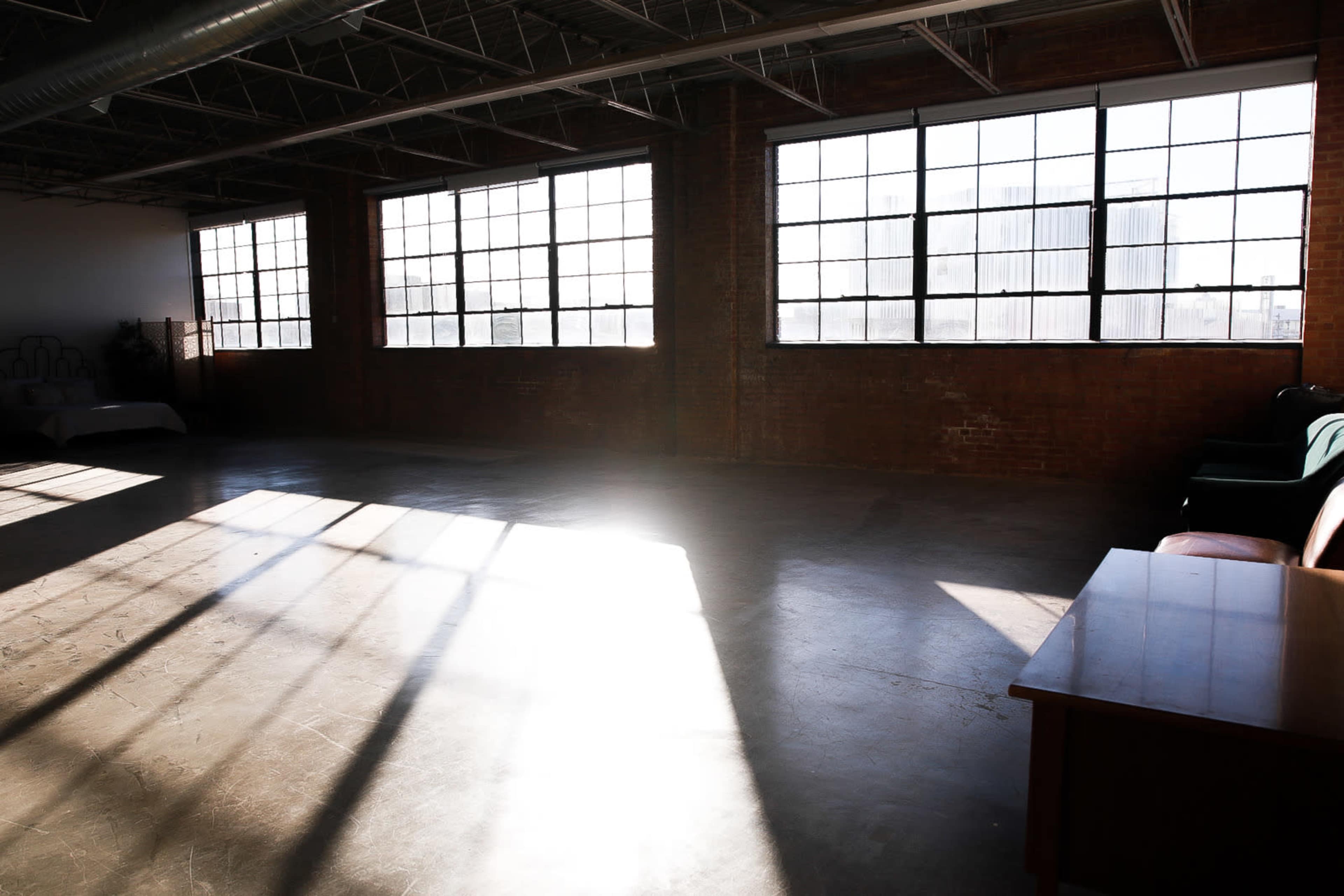 A large, empty room with concrete flooring and sunlight streaming through large industrial windows, casting shadows on the walls.