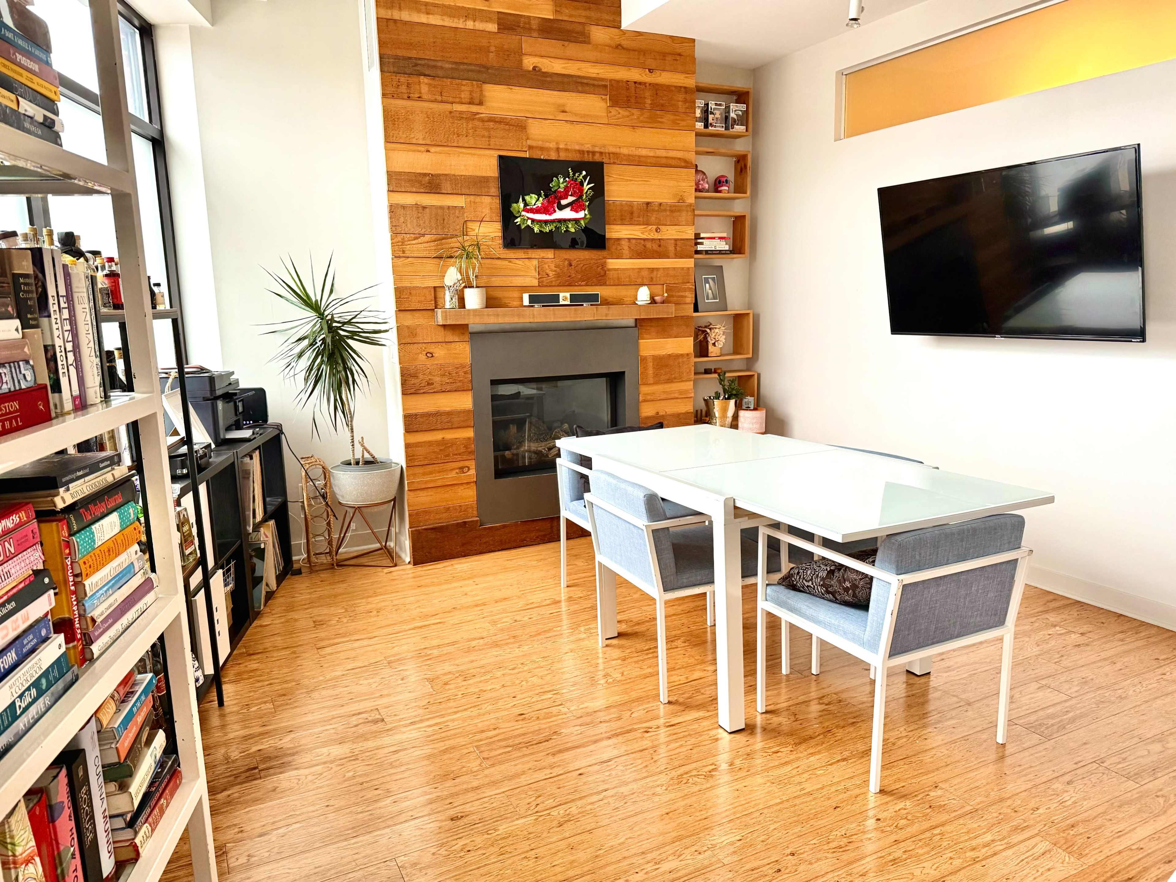 The image shows a modern living space with a wooden accent wall, a glass dining table with two chairs, a television mounted on the wall, and bookshelves filled with books.