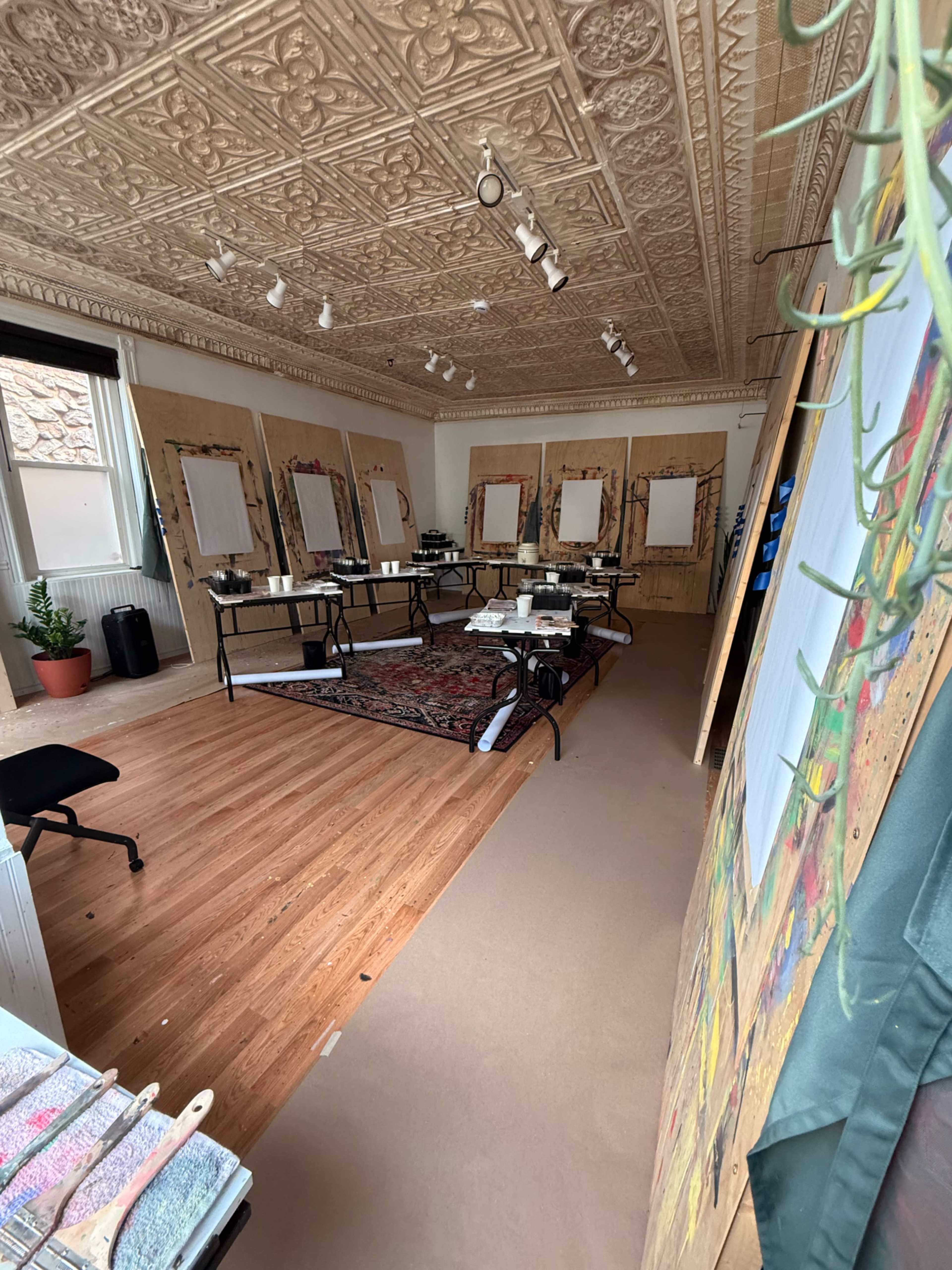 The image shows an art studio with an ornate ceiling, large empty canvases on easels, and various art supplies arranged on tables.
