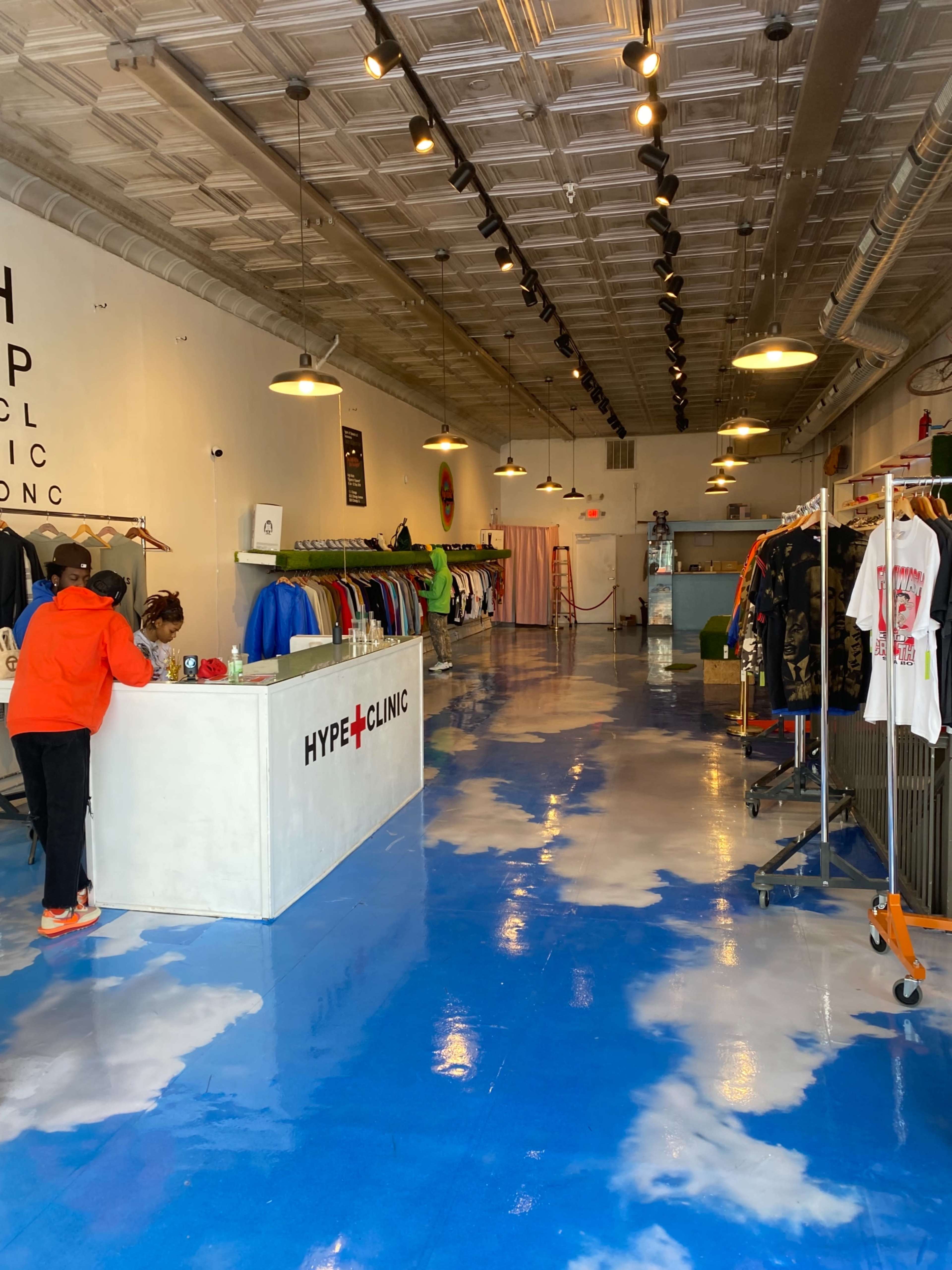 The interior of a clothing store features a check-in counter and racks displaying various apparel against a blue floor resembling a cloudy sky.