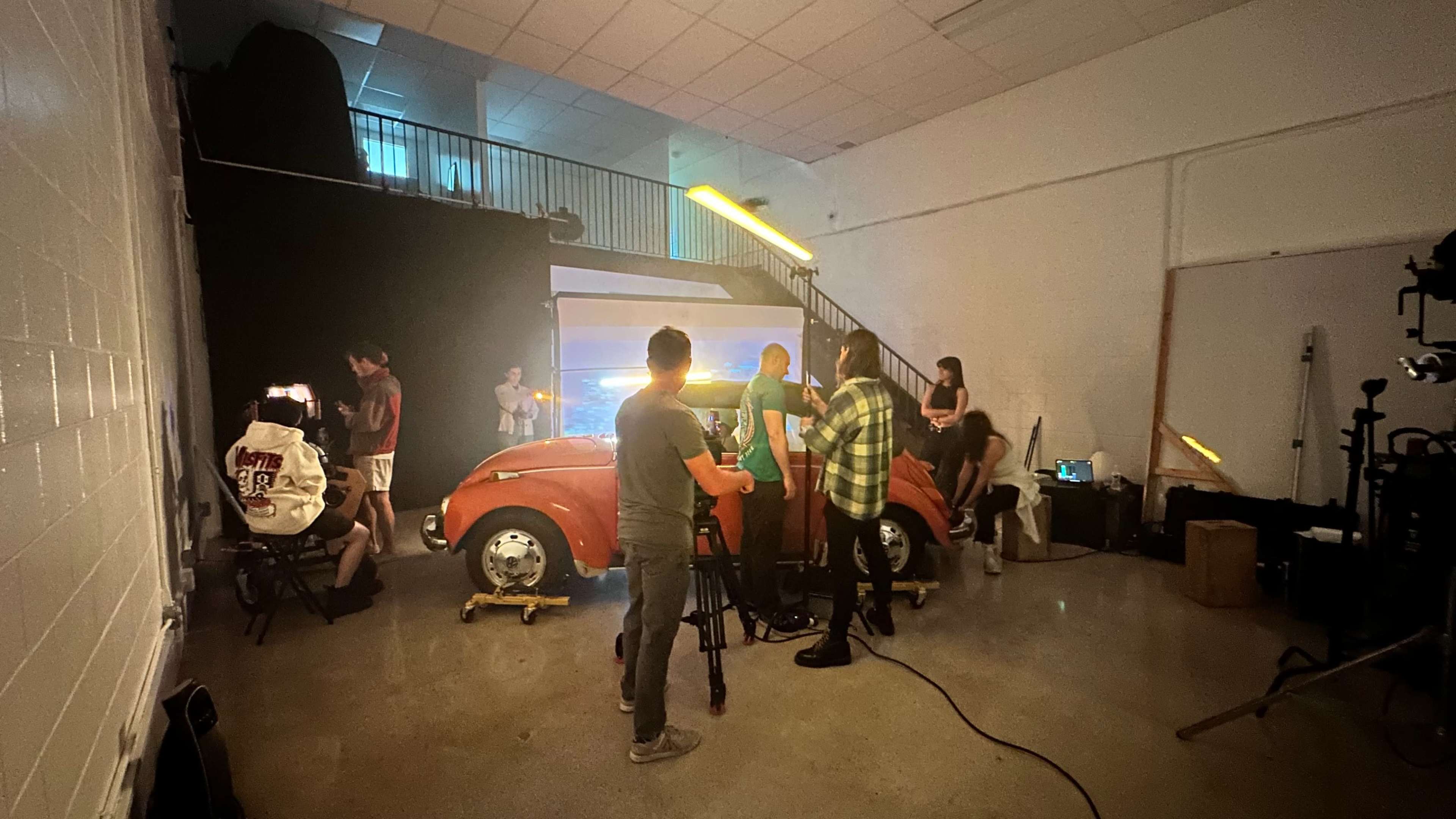 A group of people is filming a red vintage car inside a studio equipped with lighting and cameras, with additional crew members setting up in the background.