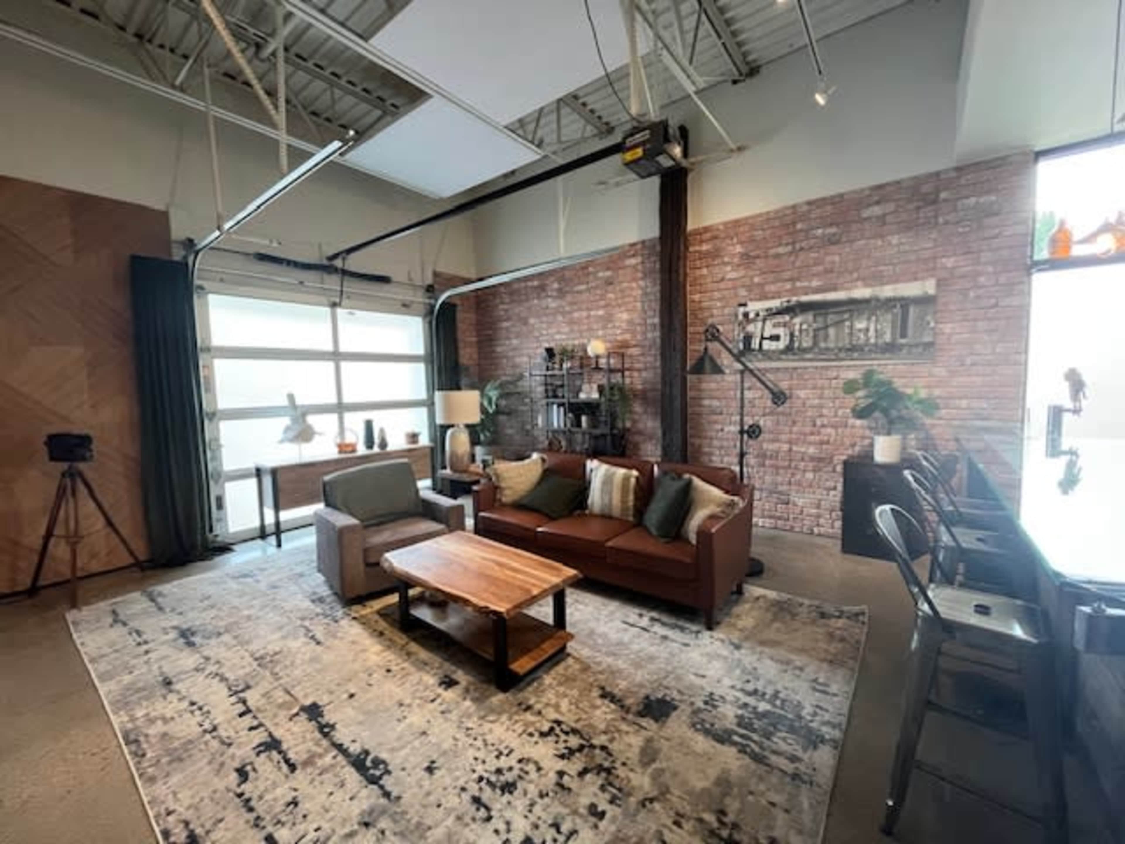 The image shows a modern living room featuring a brown leather sofa, a wooden coffee table, and a textured rug, with exposed brick walls and large windows.