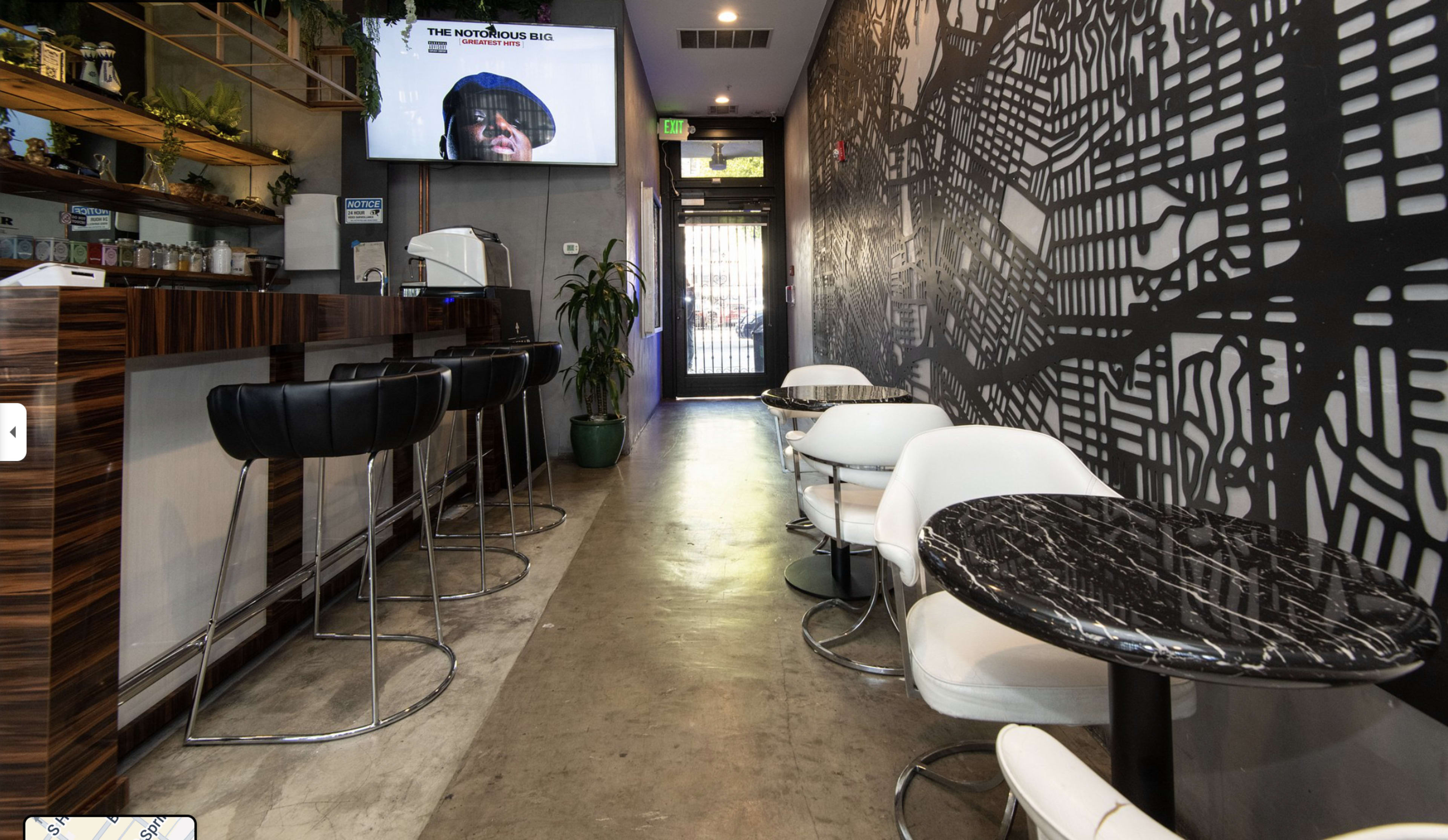 The image shows a modern café interior featuring a bar with stools on the left, small round tables with white chairs on the right, and a wall adorned with a large abstract mural.