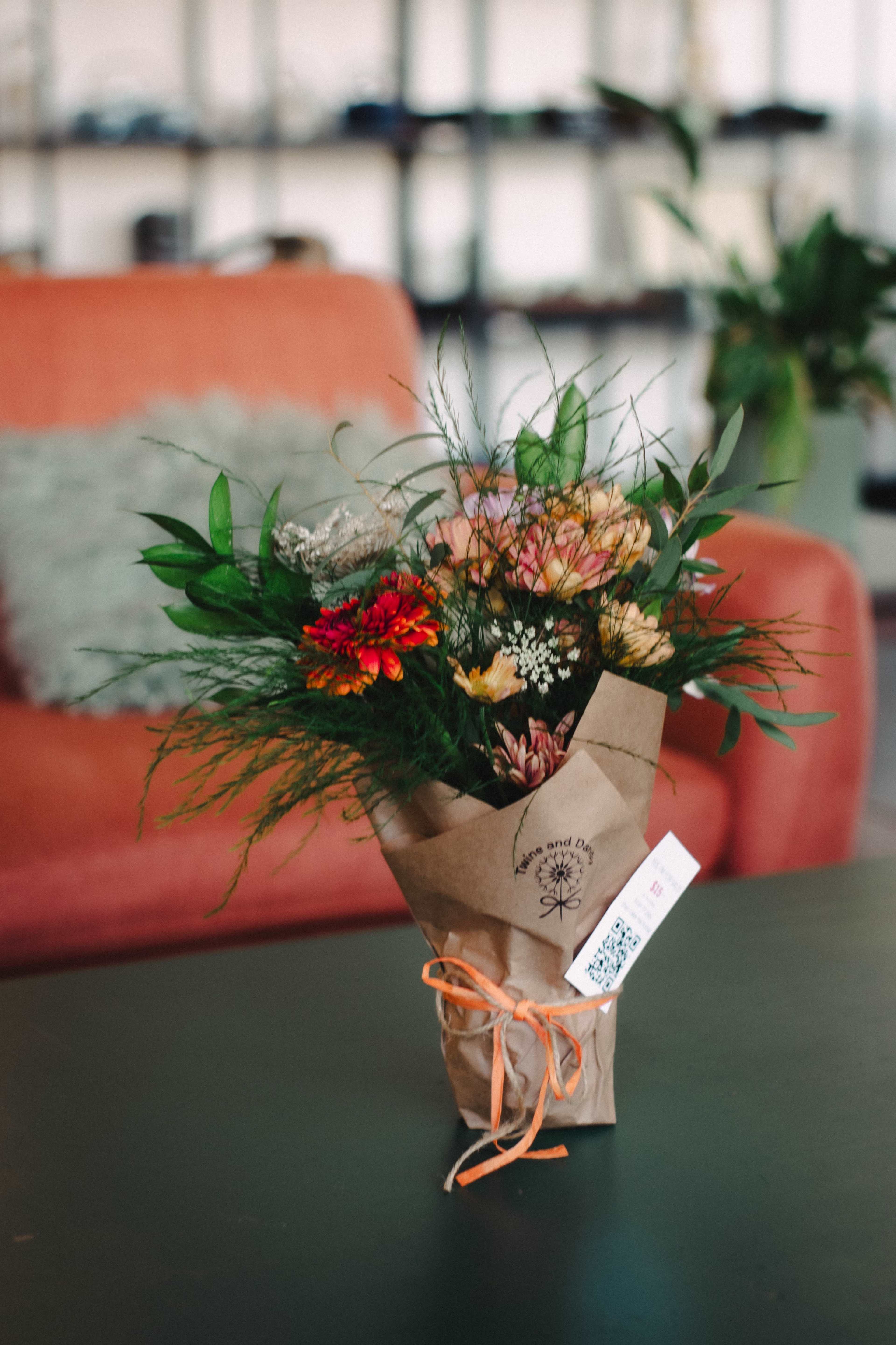 A bouquet of assorted flowers, wrapped in paper and tied with an orange ribbon, sits on a black table in a room with a red couch in the background.