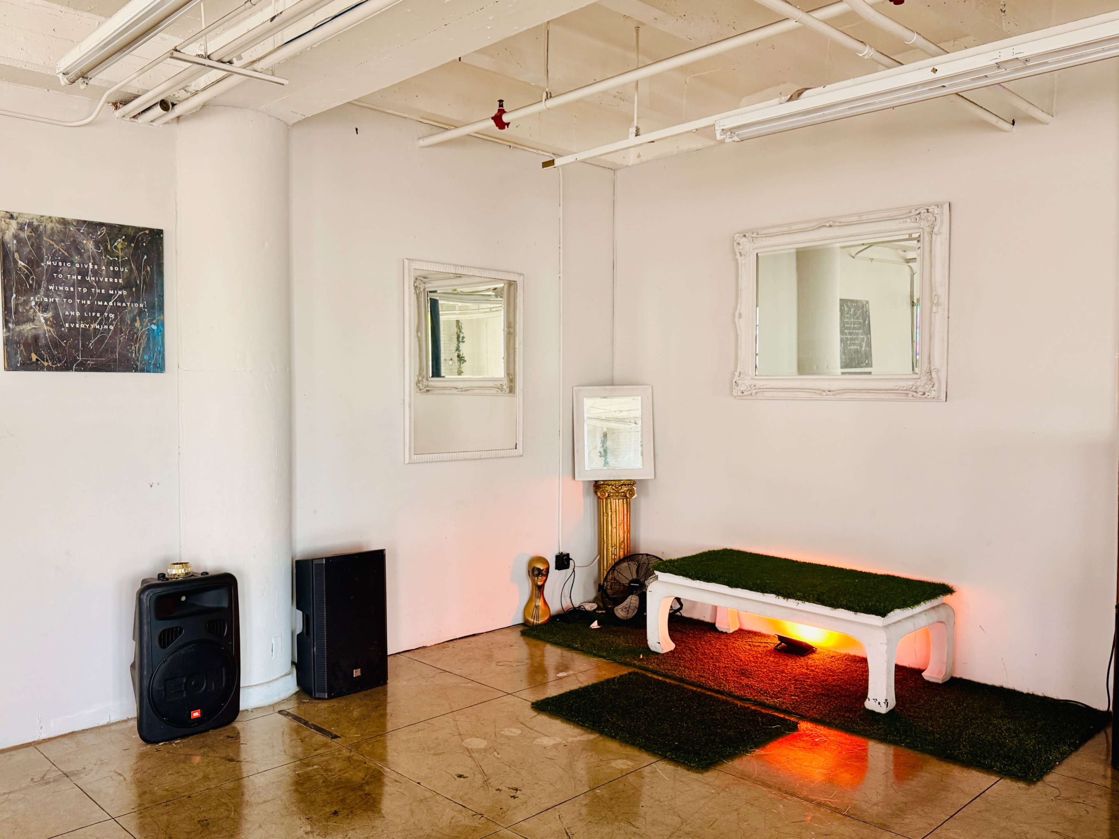 The image shows a minimalist interior space with a white wall, a green rug, a white bench, mirrors, and speakers.
