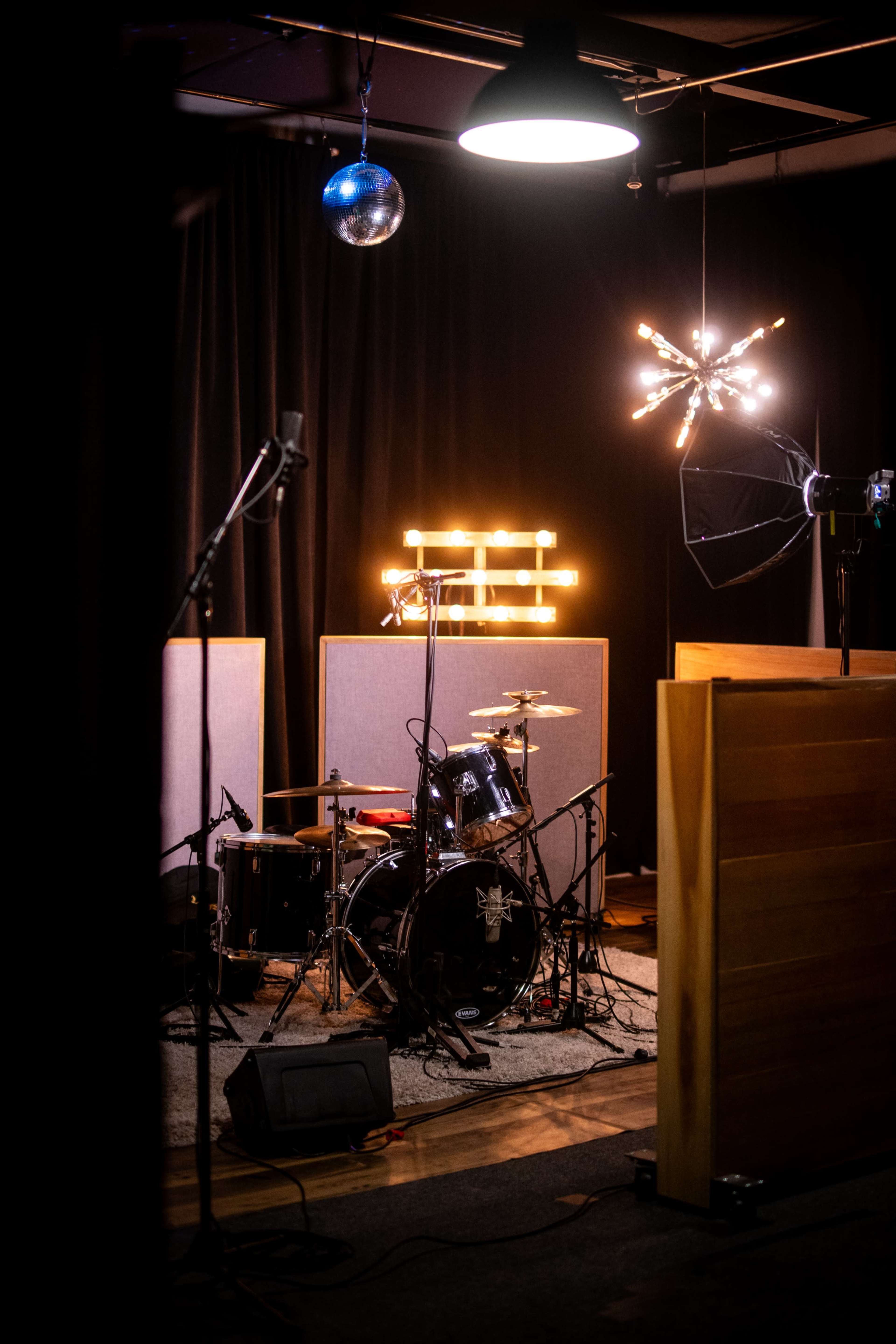The image shows a music studio setup featuring a drum kit in front of soundproofing panels, with decorative lighting overhead.