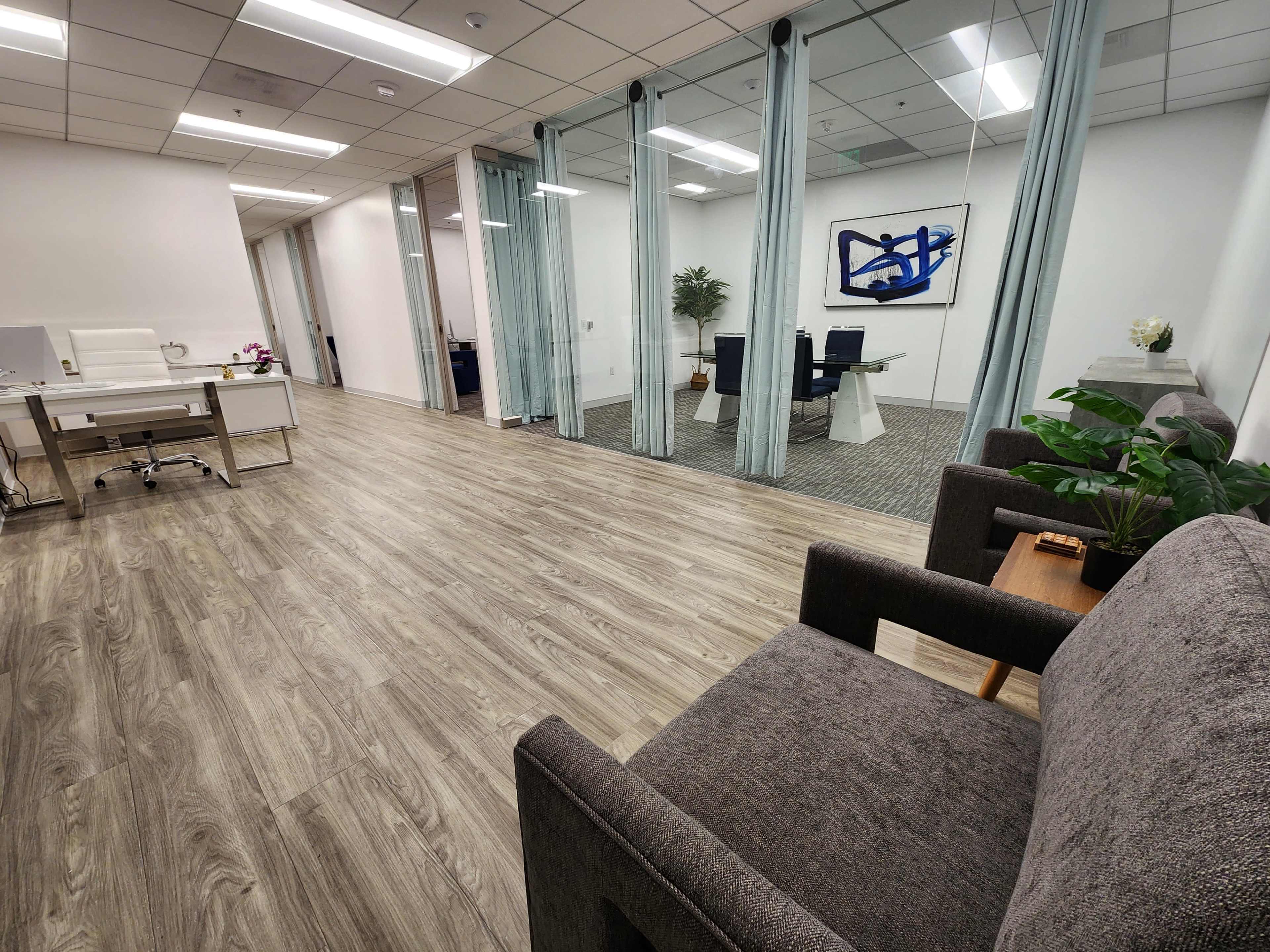 The image shows a modern office space featuring a wood floor, a seating area with gray chairs, and glass partitions separating meeting areas.