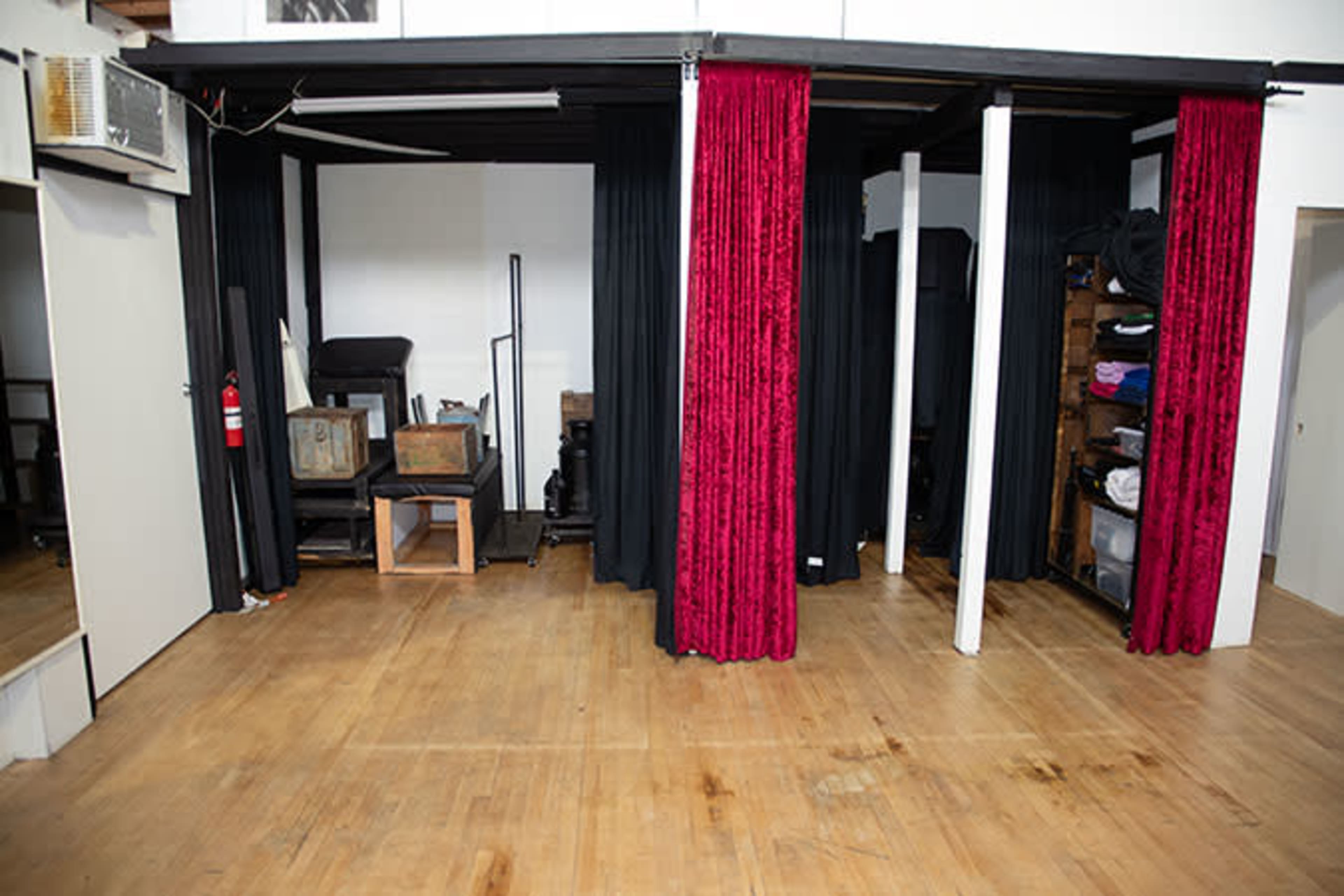The image shows an empty room with wooden floors, draped black curtains, and red curtains separating different areas, along with some furniture and storage units against the walls.