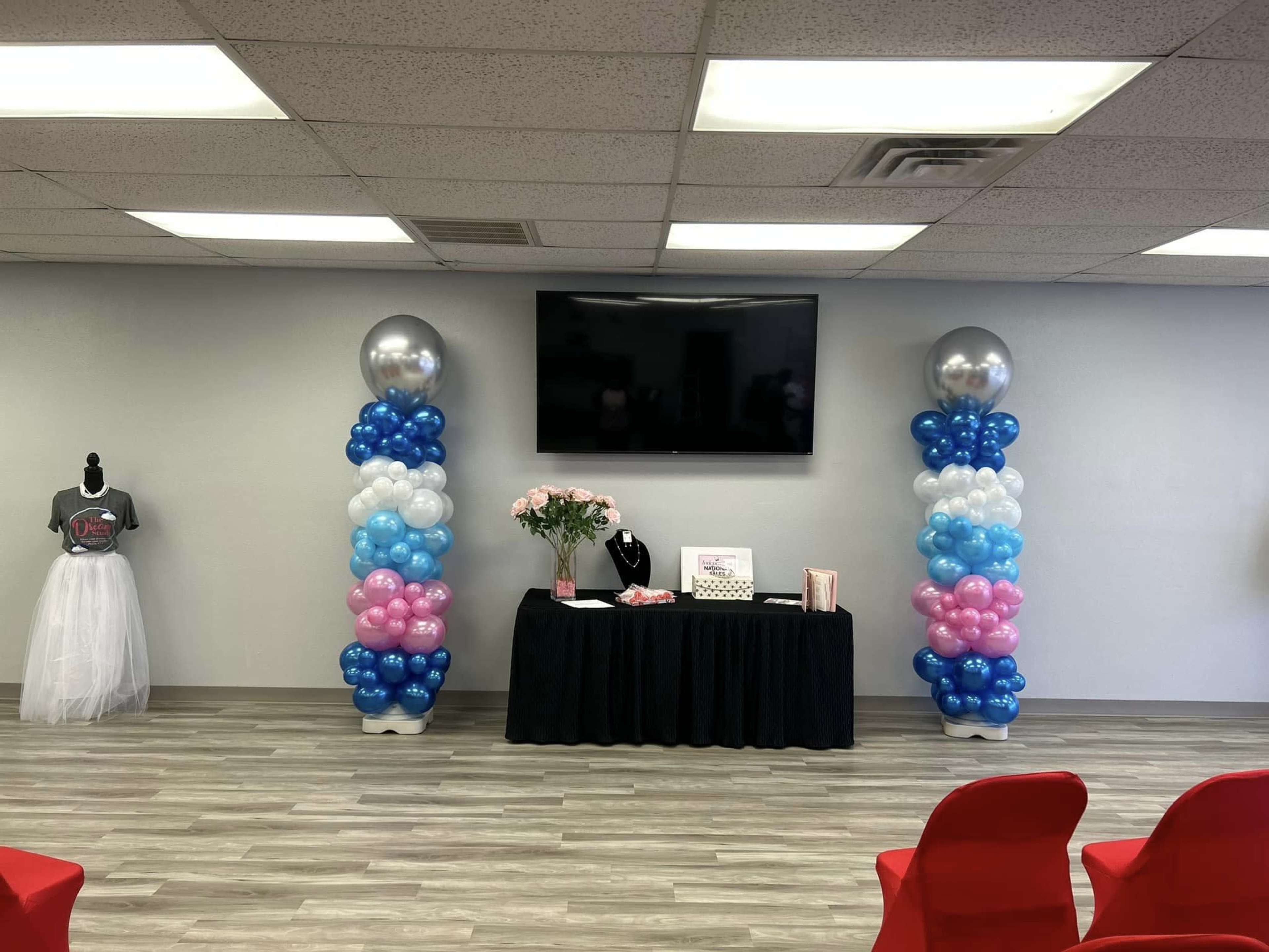 A decorated room with two balloon columns in blue, white, and pink, a black table displaying flowers and items, and a television mounted on the wall.