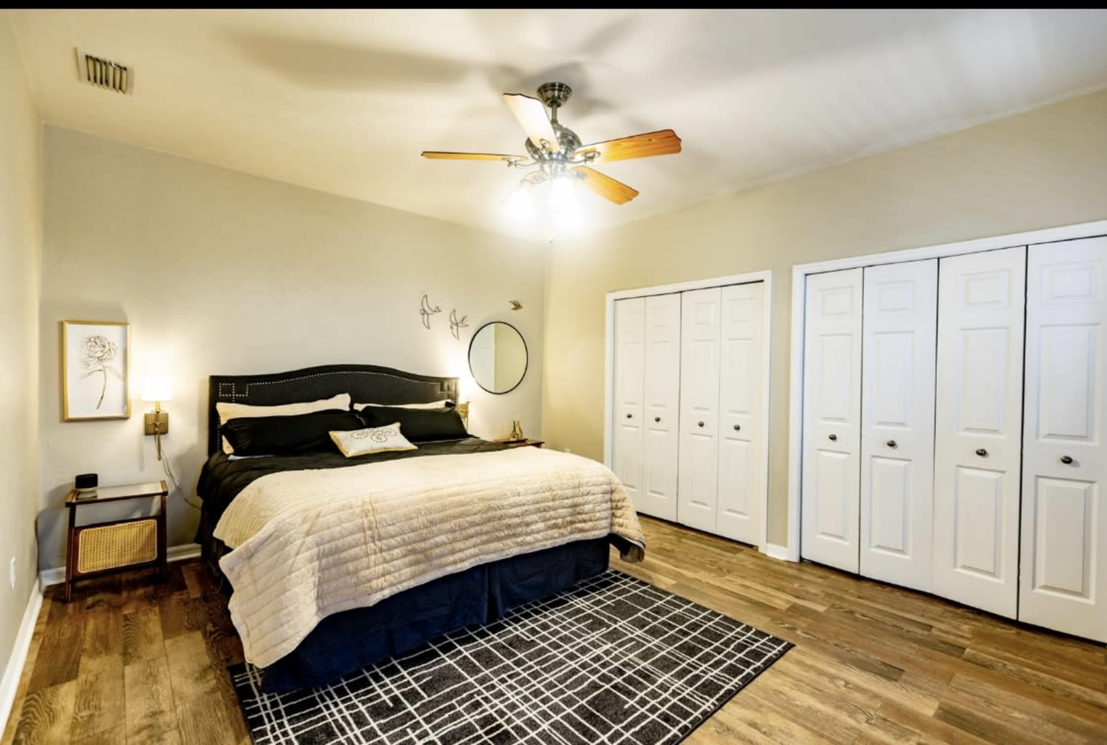 The image shows a well-decorated bedroom featuring a large bed with a black and beige color scheme, a ceiling fan, and two white closet doors.