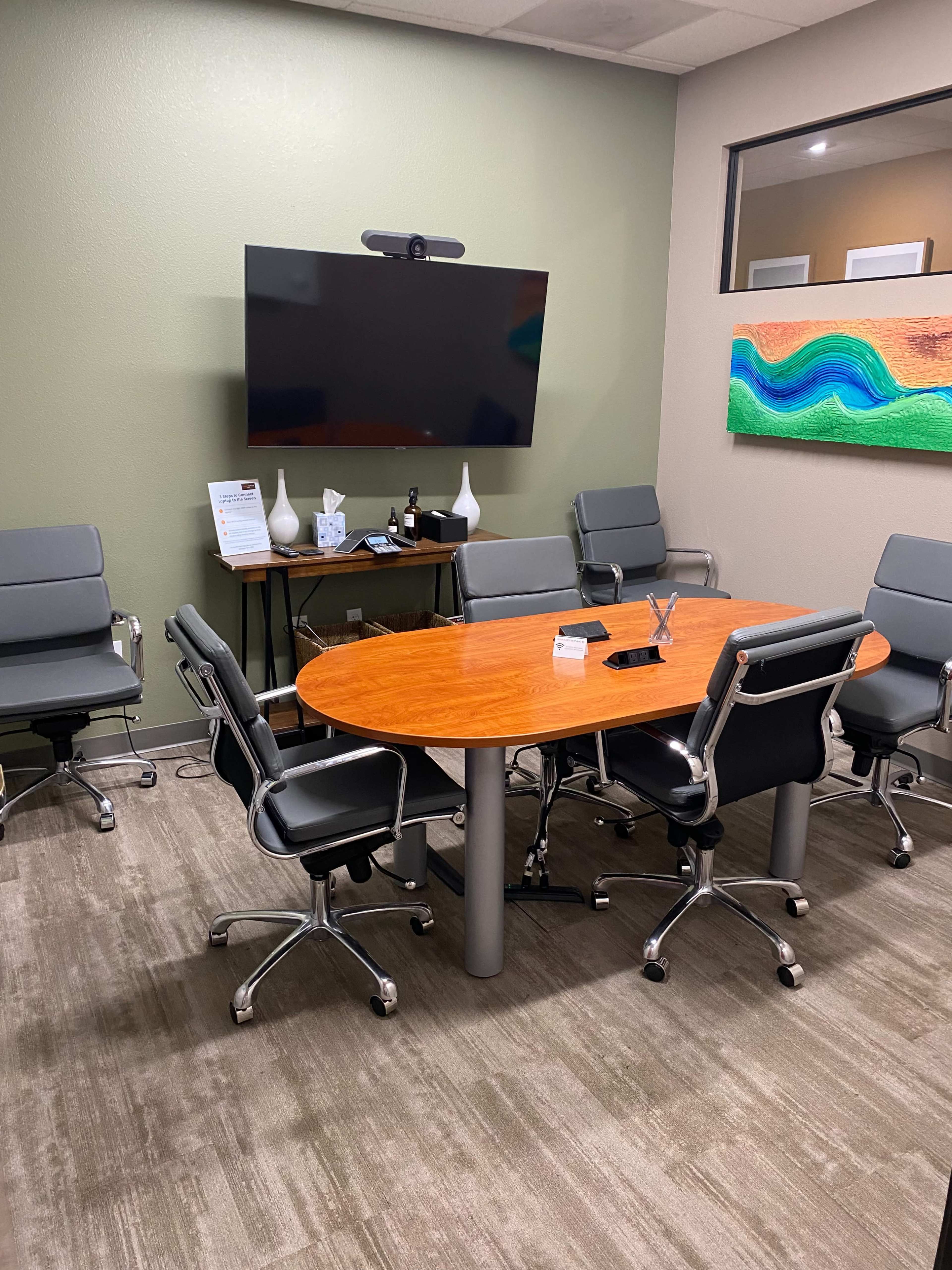 A conference room with a wooden oval table surrounded by six gray chairs, a large wall-mounted TV, and an abstract painting on the wall.