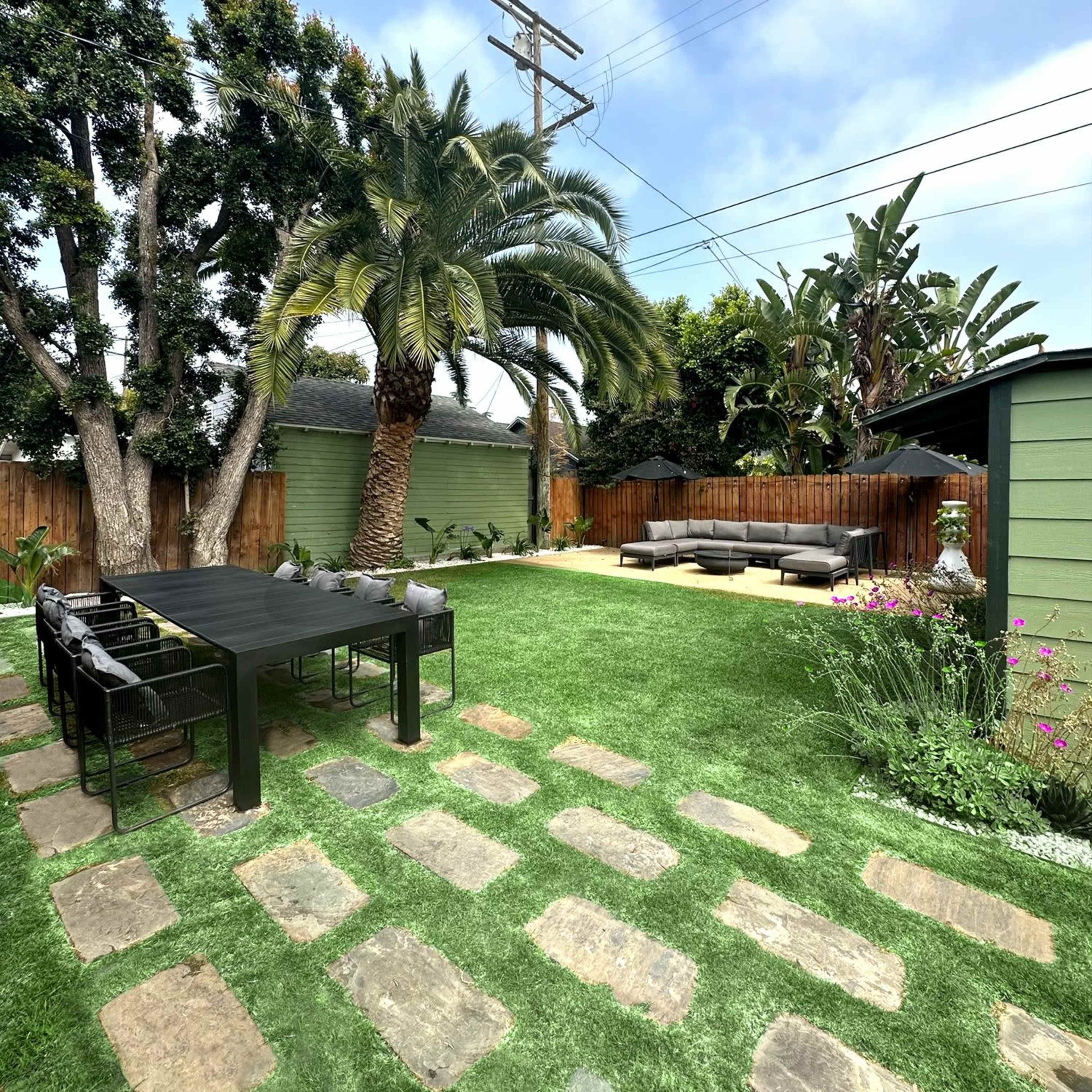 A landscaped backyard features a stone pathway, a black dining table, palm trees, and a seating area with outdoor furniture.