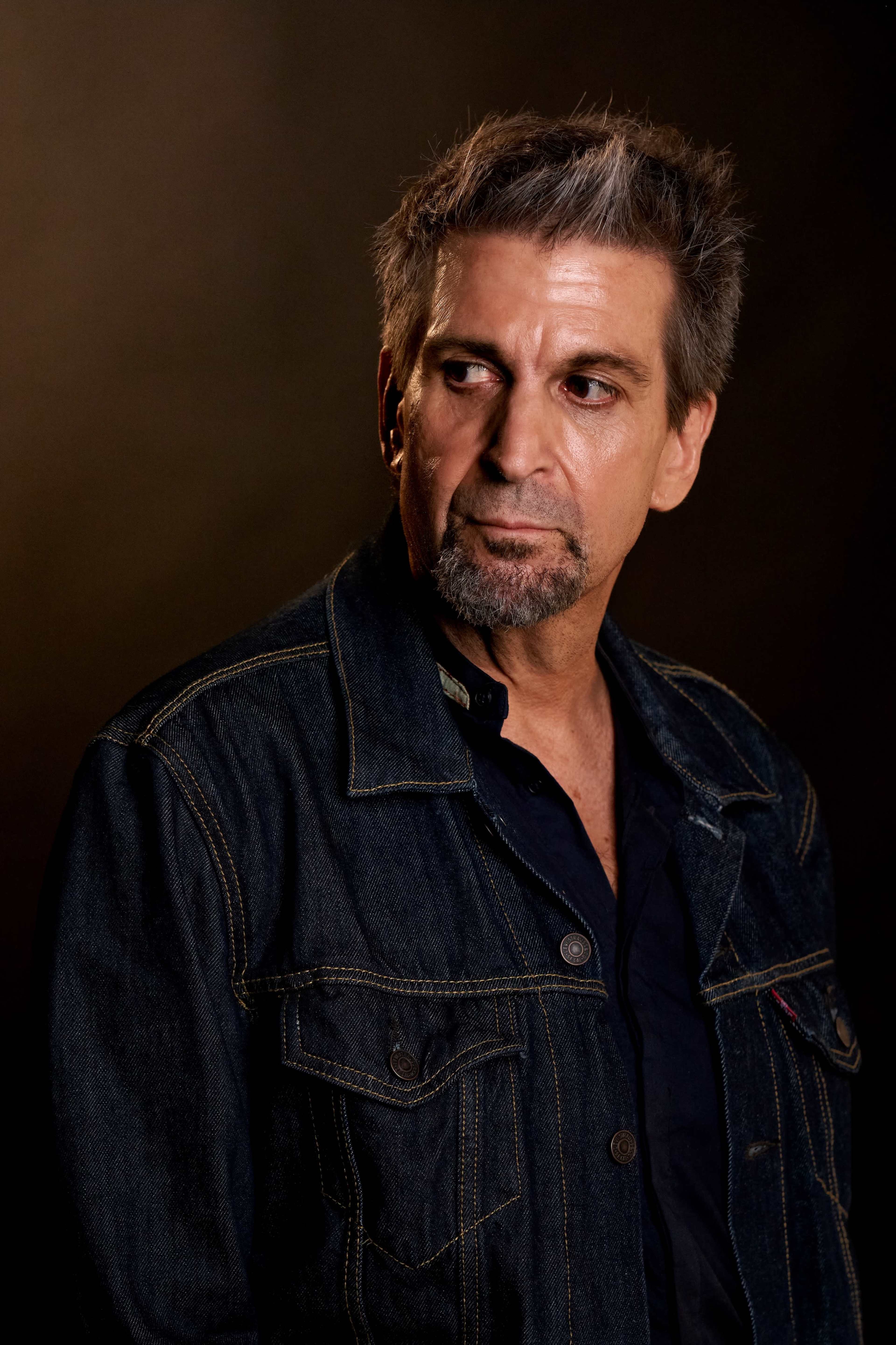 A man with gray-streaked hair wears a denim jacket and looks off to the side against a dark background.