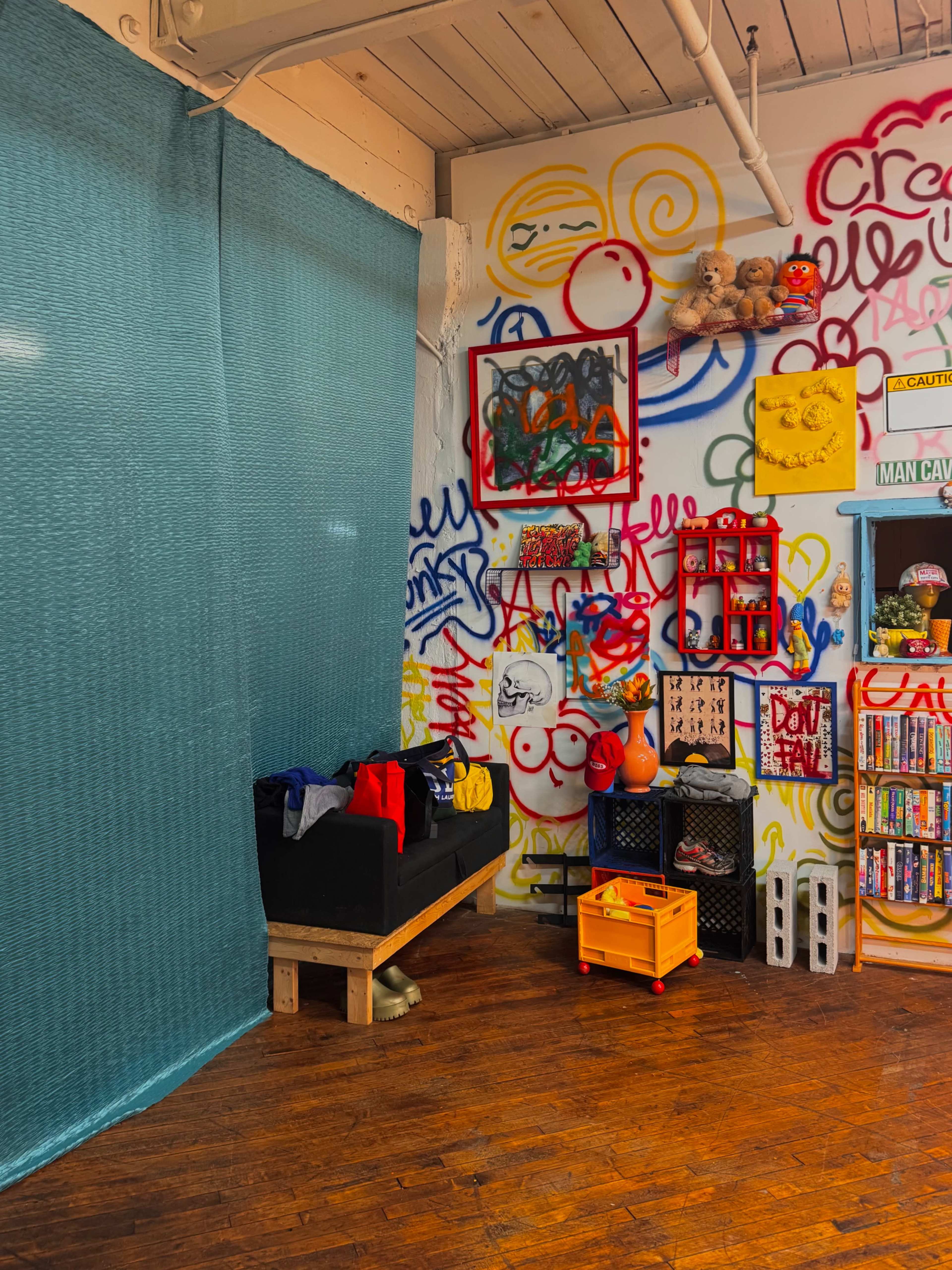 The image features a colorful room with a graffiti-covered wall, a blue curtain on one side, and a variety of decorative items and furniture, including a black bench and colorful storage crates.