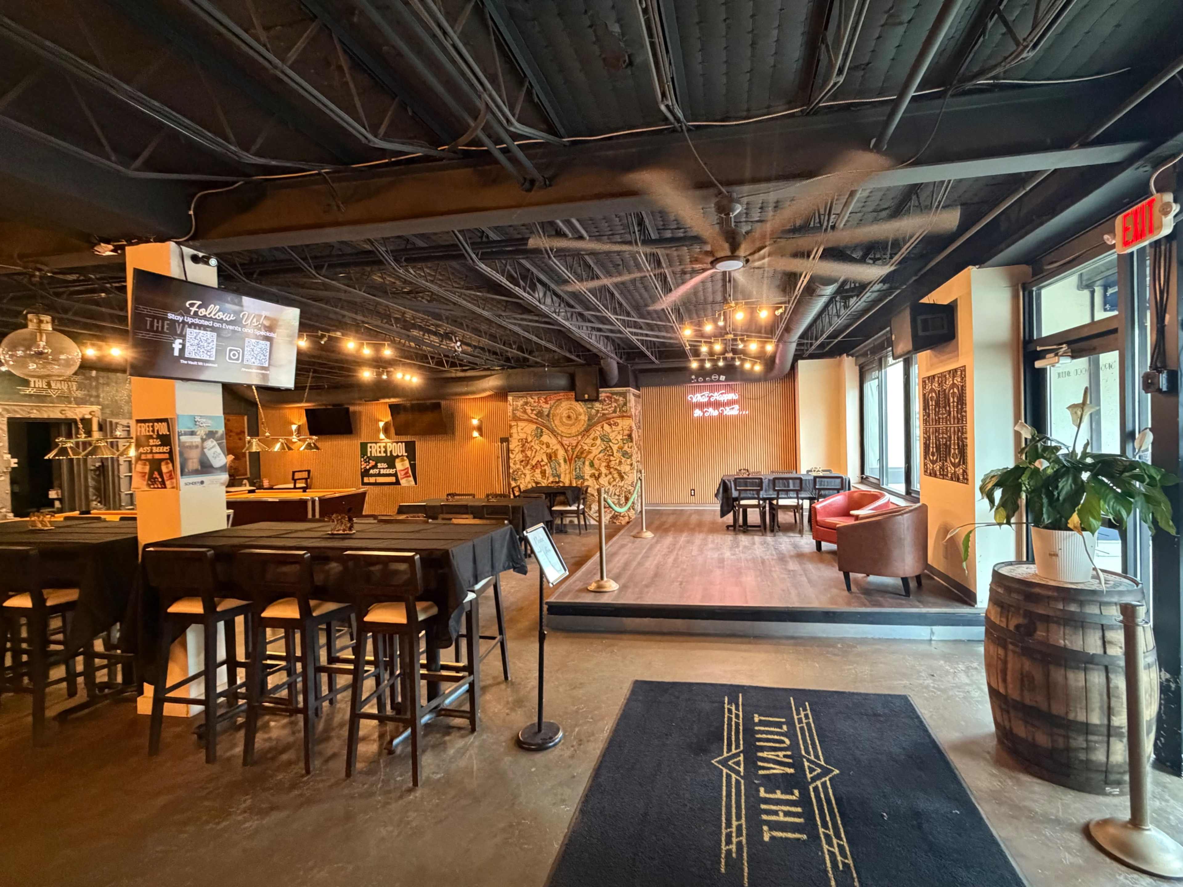 The image shows the interior of a dimly lit bar or lounge area featuring a mix of seating arrangements, decorative wall art, and a welcoming entrance.