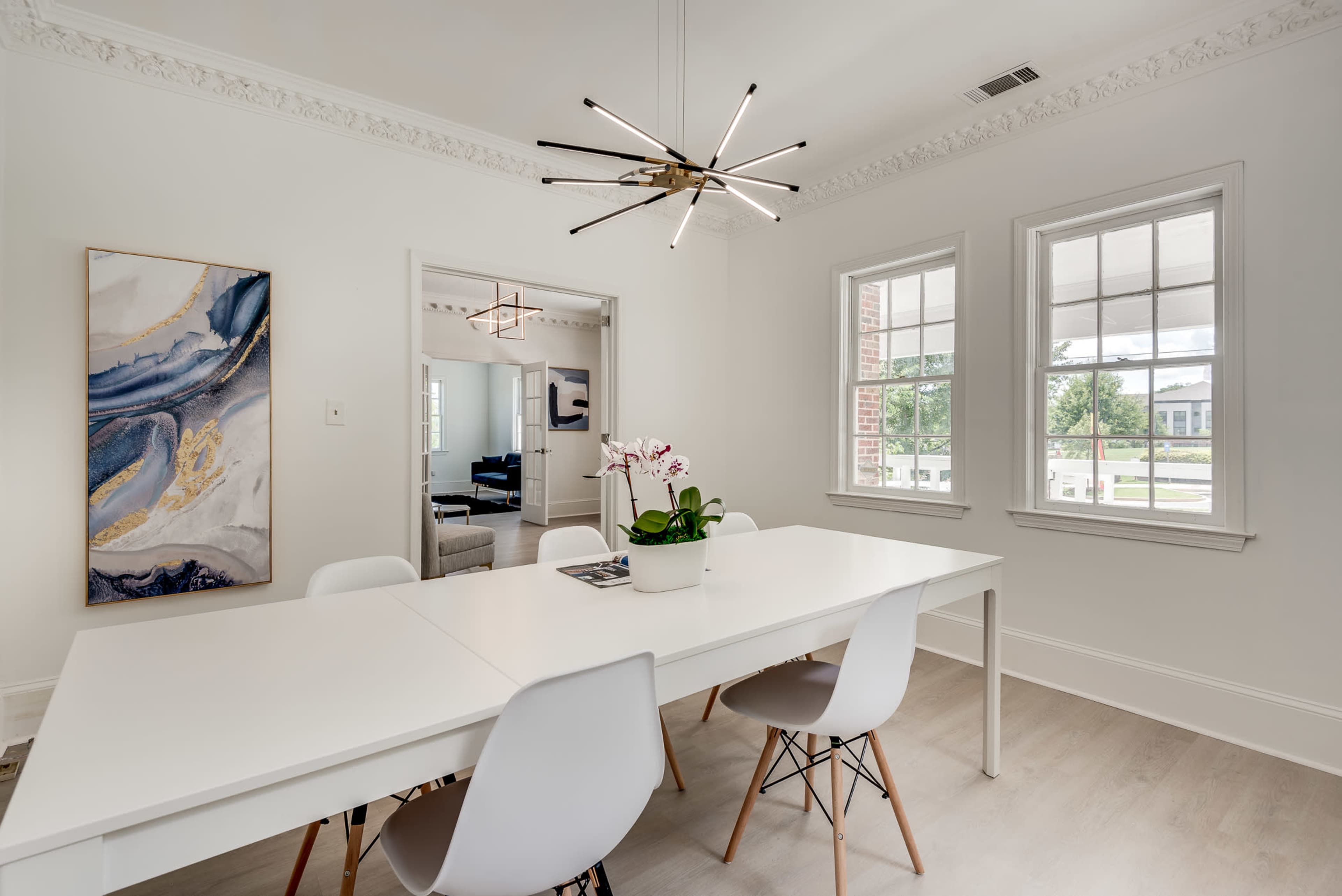 A modern dining area features a long white table surrounded by five light chairs, with a chandelier above and large windows on the side.