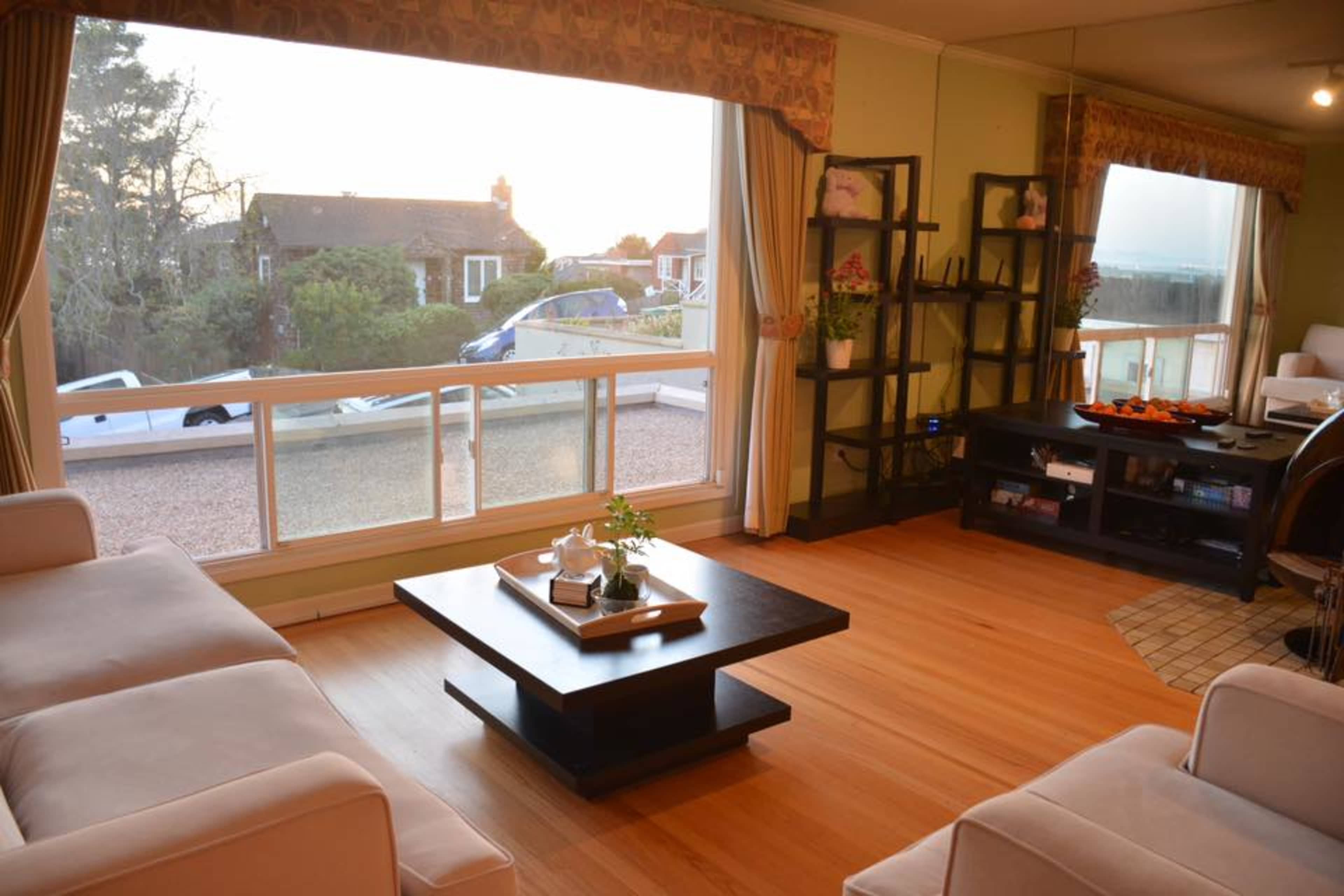 The image shows a sunlit living room with two sofas, a coffee table, and a large window offering a view of the outdoors.