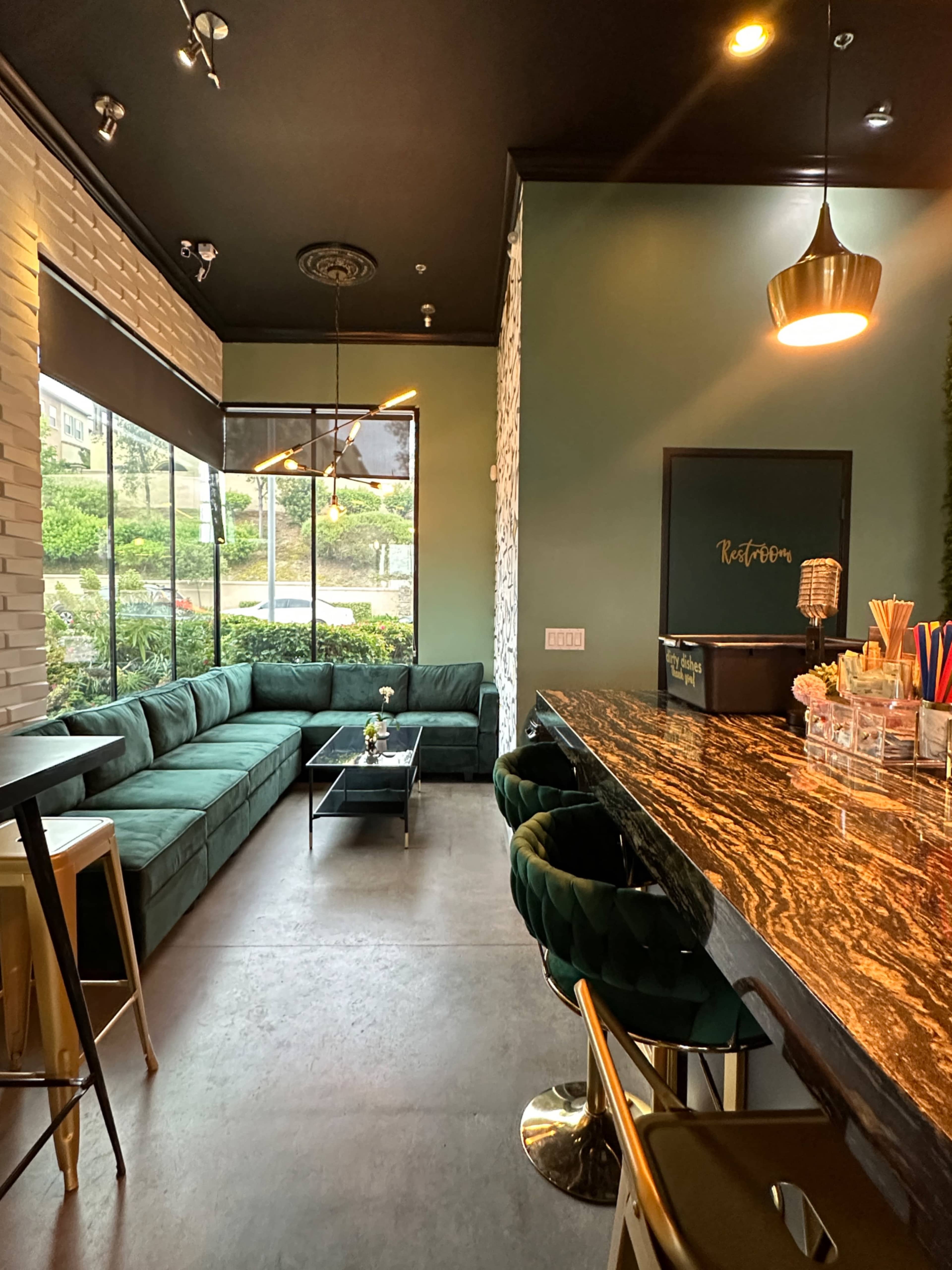 The image shows a modern cafe interior featuring a green velvet seating area, a marble countertop bar, and large windows overlooking an outdoor space.