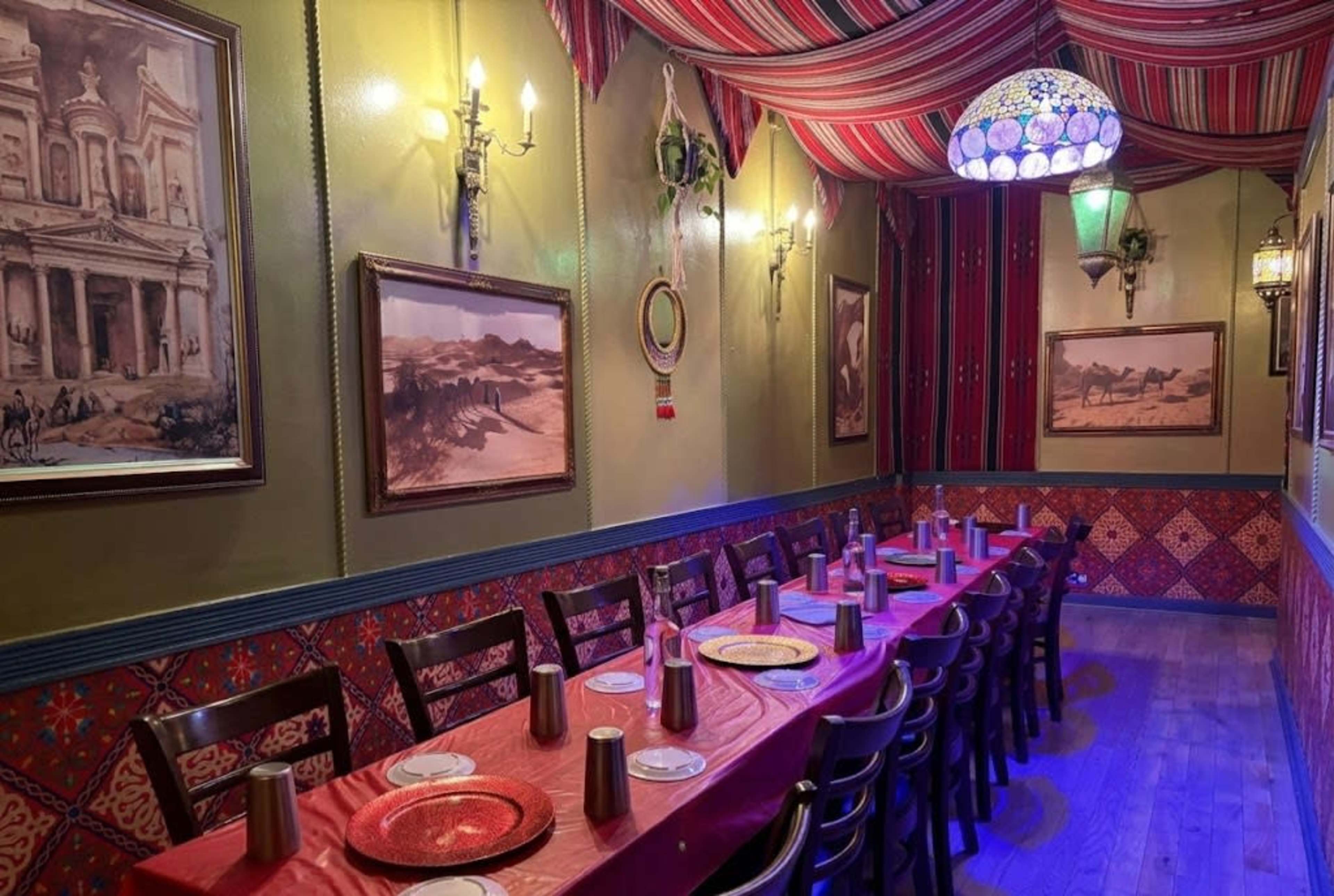 The image depicts a restaurant interior featuring a long table set with plates and decorative cups, surrounded by patterned walls and paintings.