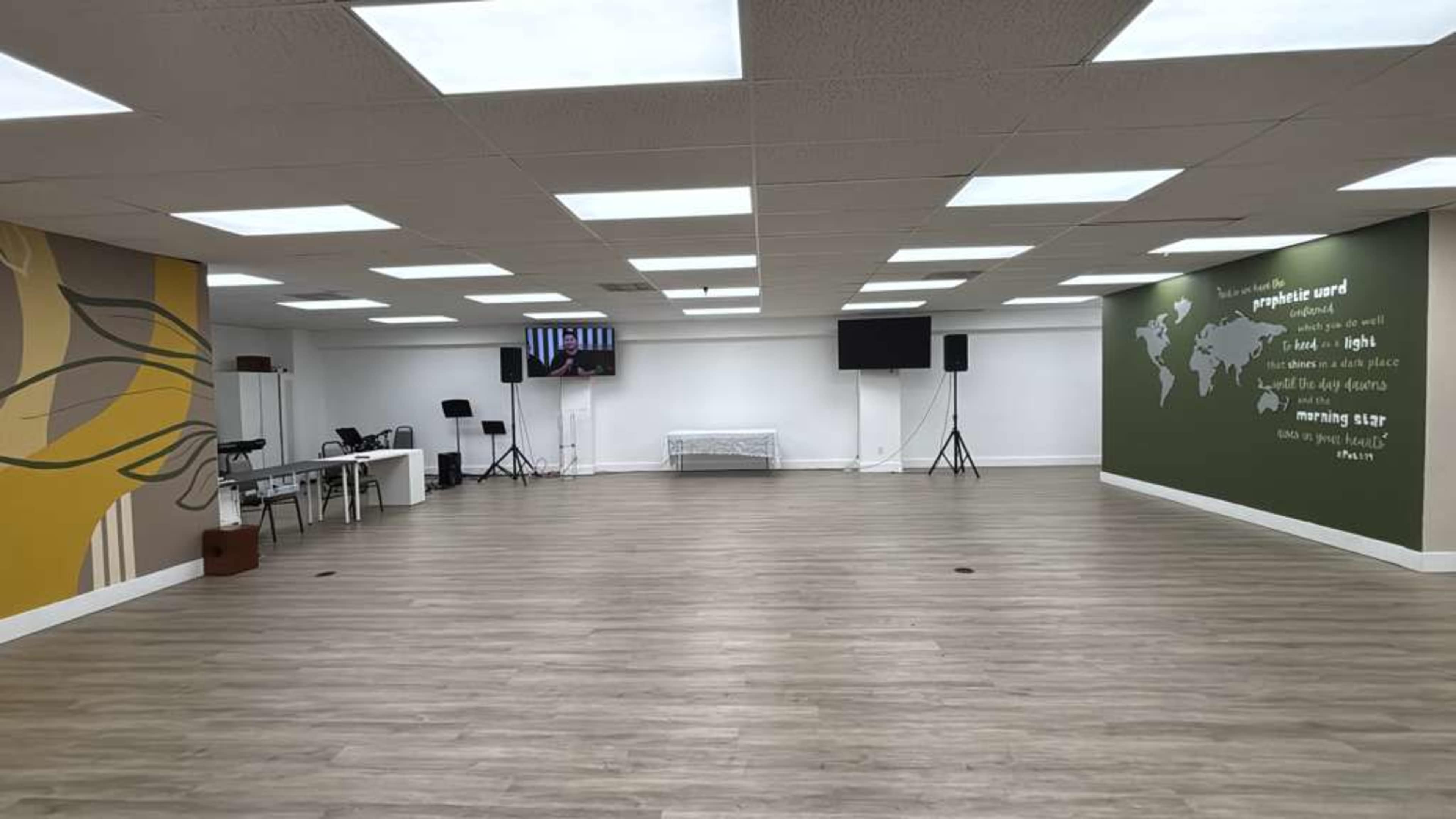 The image shows a spacious, well-lit interior room with wooden flooring, featuring a large mural on one wall and two screens on stands.