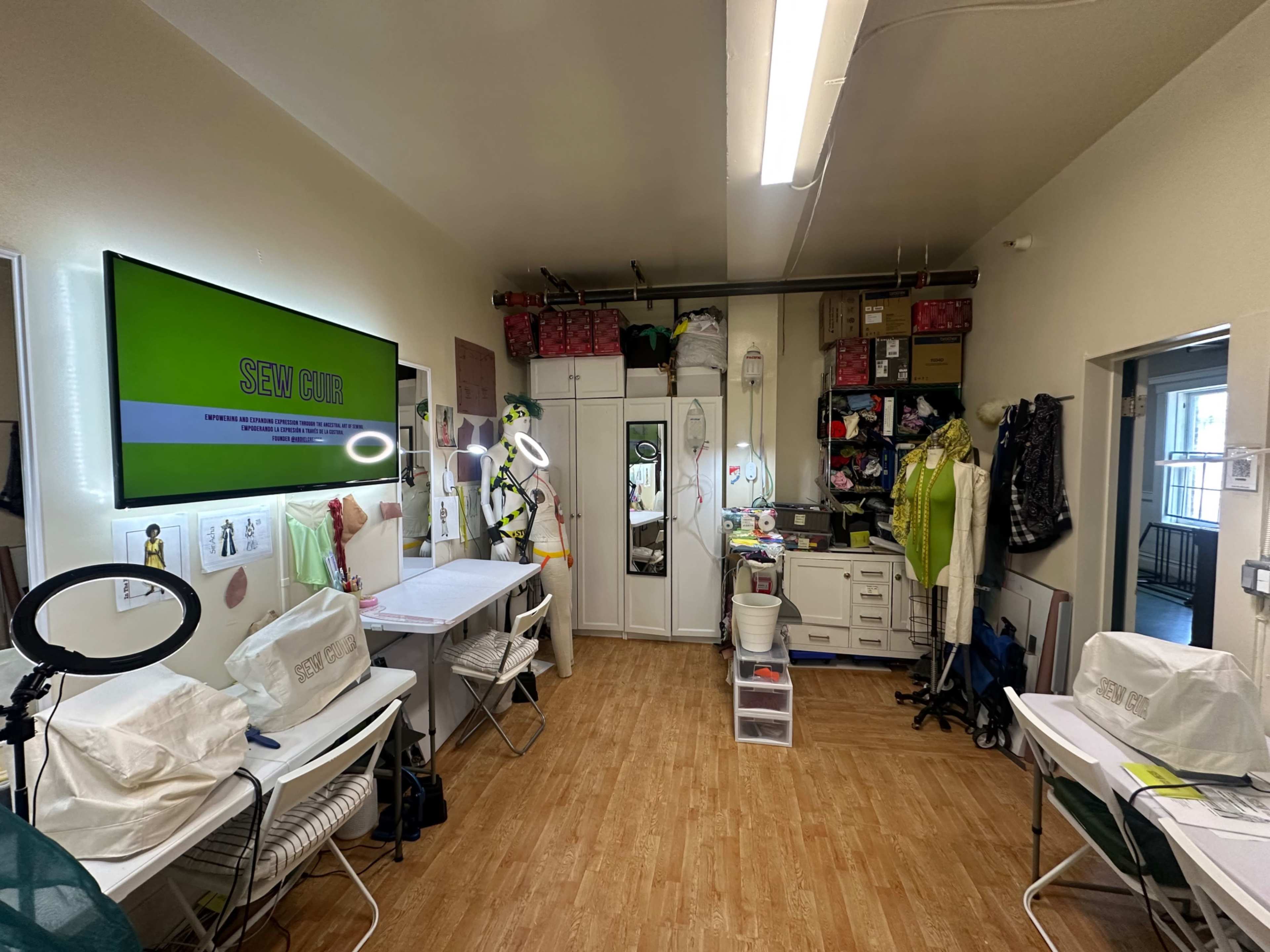 The image shows a sewing studio with several tables, organized storage, and a large screen displaying the words "SEW CUT."