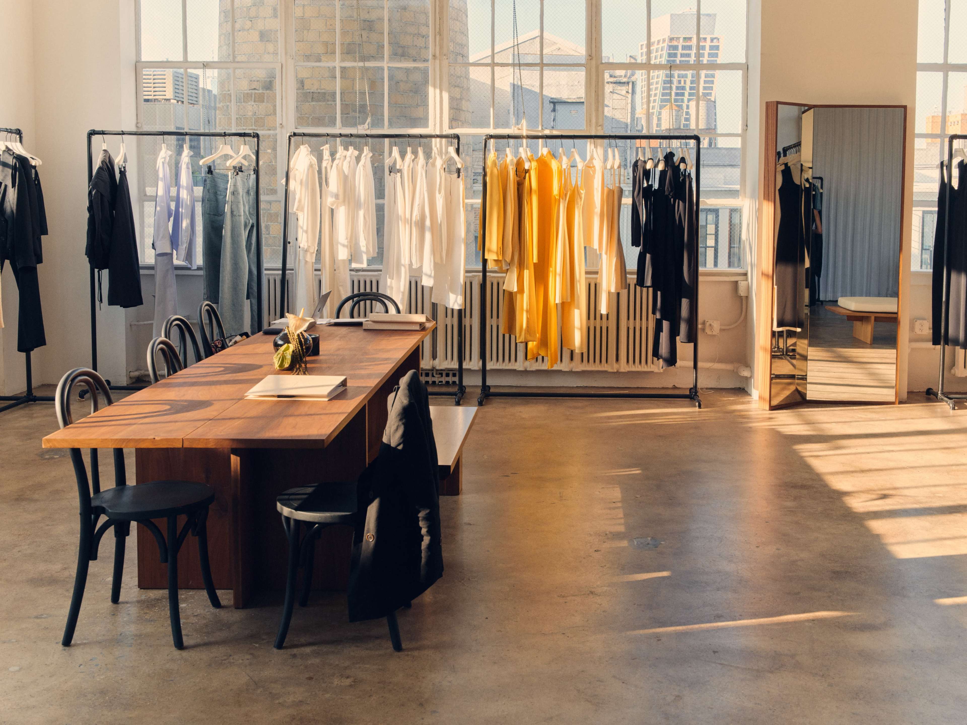The image shows a spacious clothing showroom with a large wooden table and racks of garments in various colors arranged against the walls.