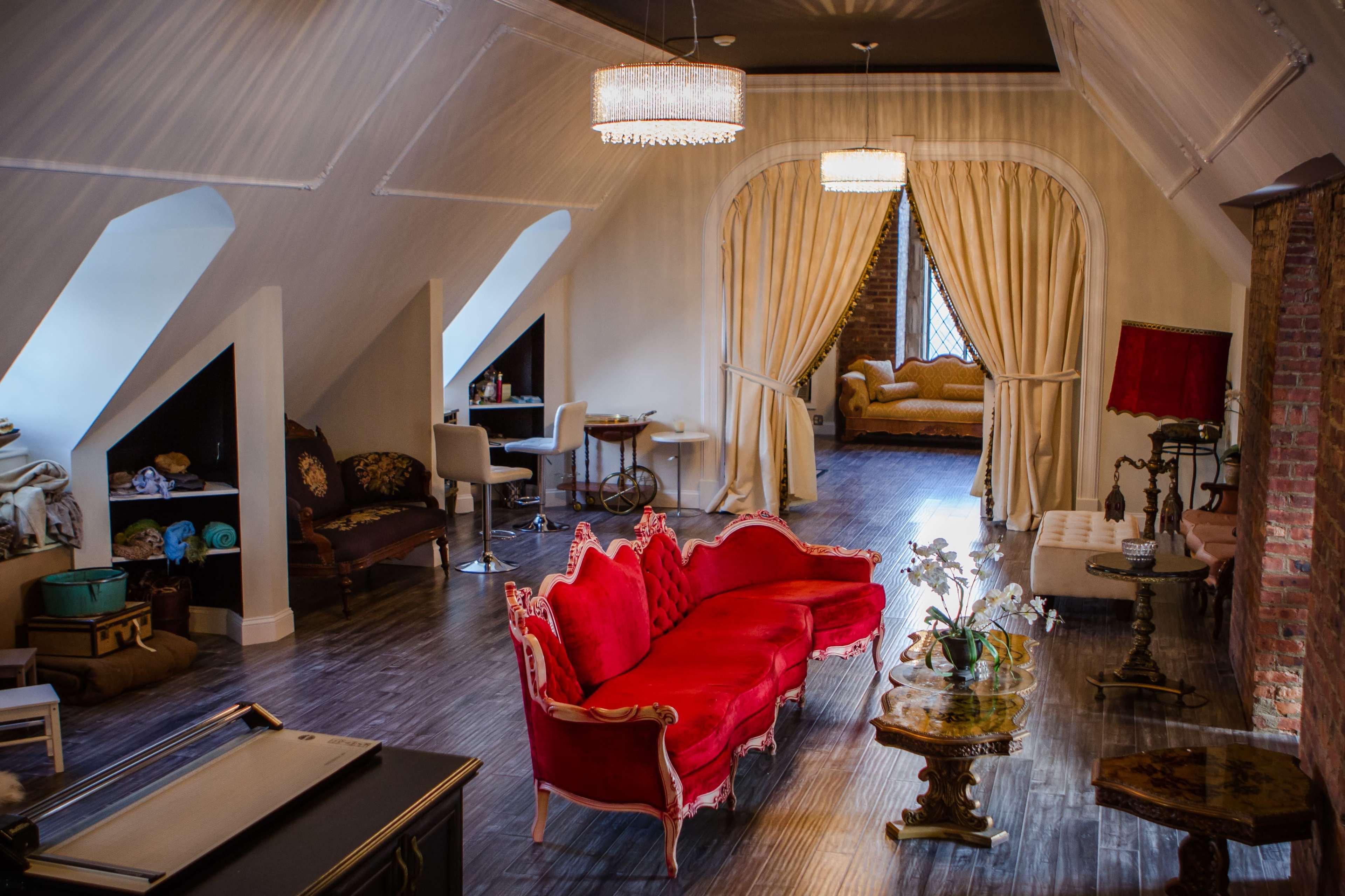The scene depicts a well-decorated attic space featuring a red upholstered sofa, elegant lighting, and a cozy seating area with a view of a bed behind decorative curtains.