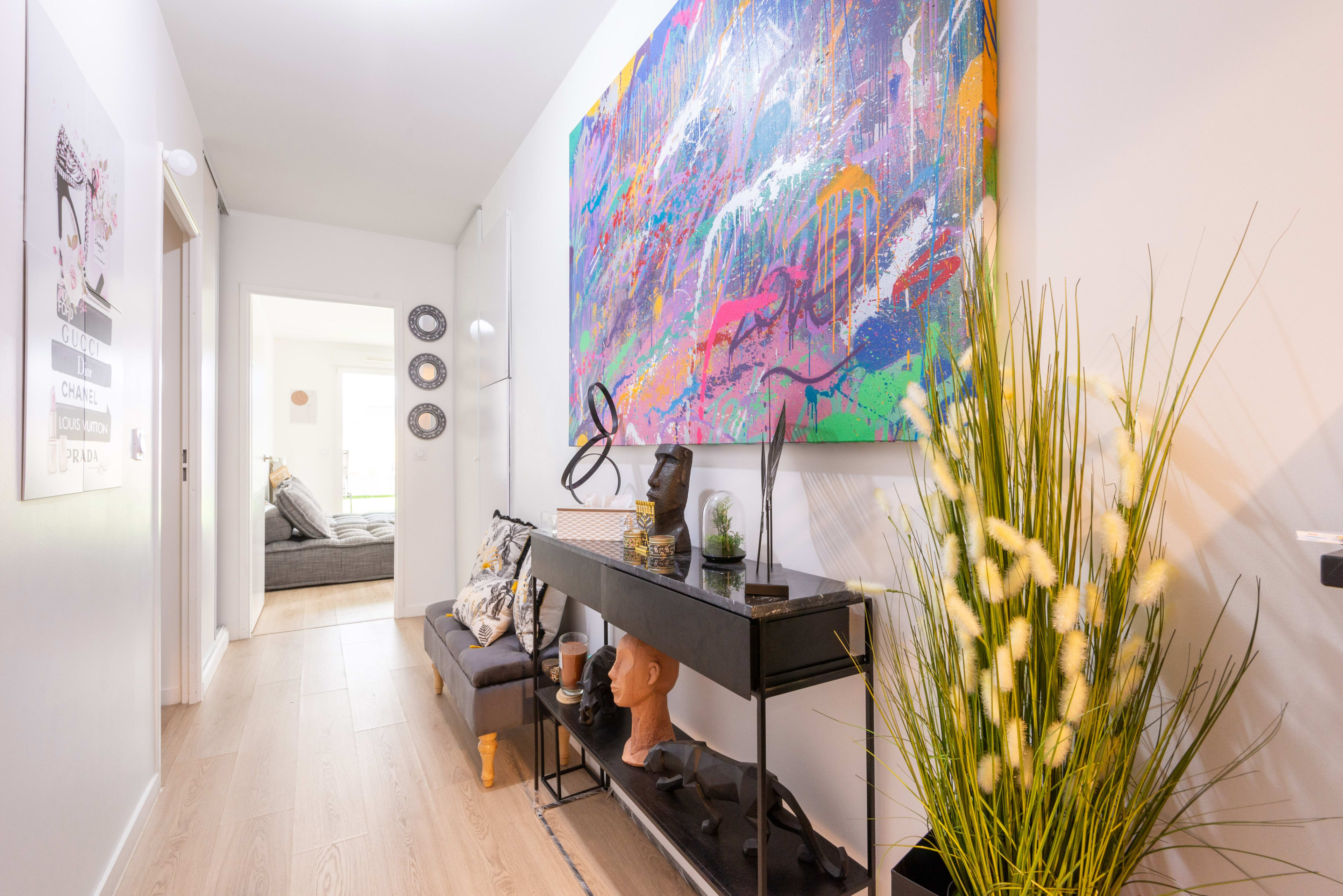 A hallway features a colorful abstract painting above a sleek console table adorned with decorative items and potted plants, leading to a well-lit room beyond.
