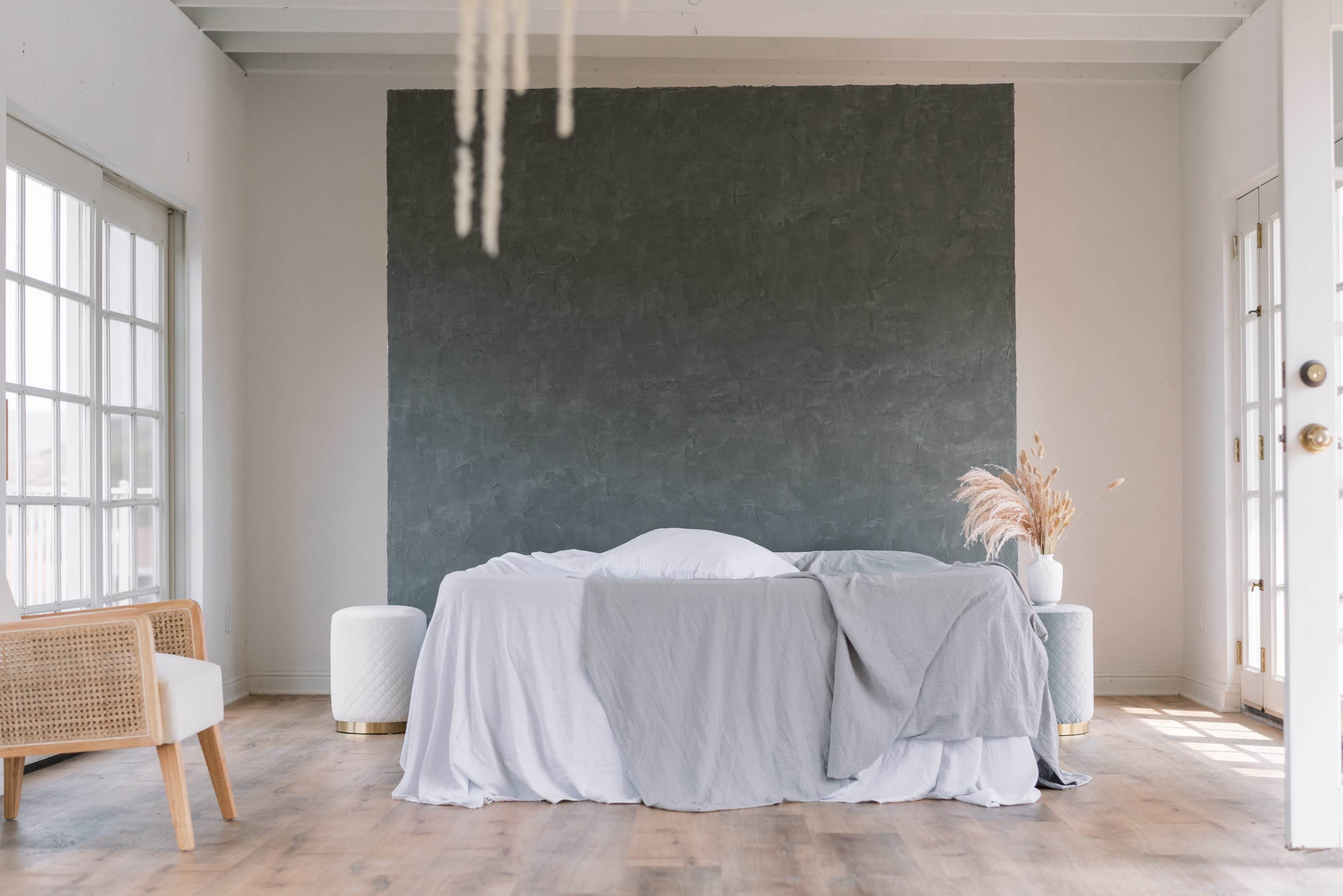 A bed with gray linens is centered in a minimally decorated room featuring large windows and a textured dark wall.